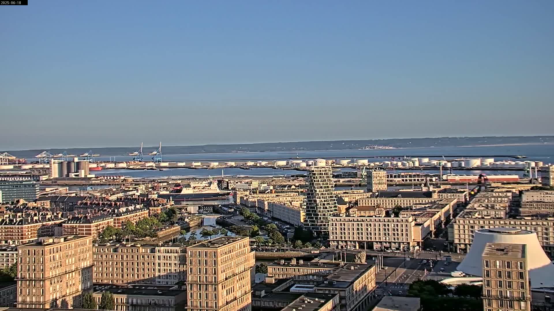 A sprawling cityscape with numerous buildings and a harbor filled with industrial structures and ships, under a clear, sunny sky.