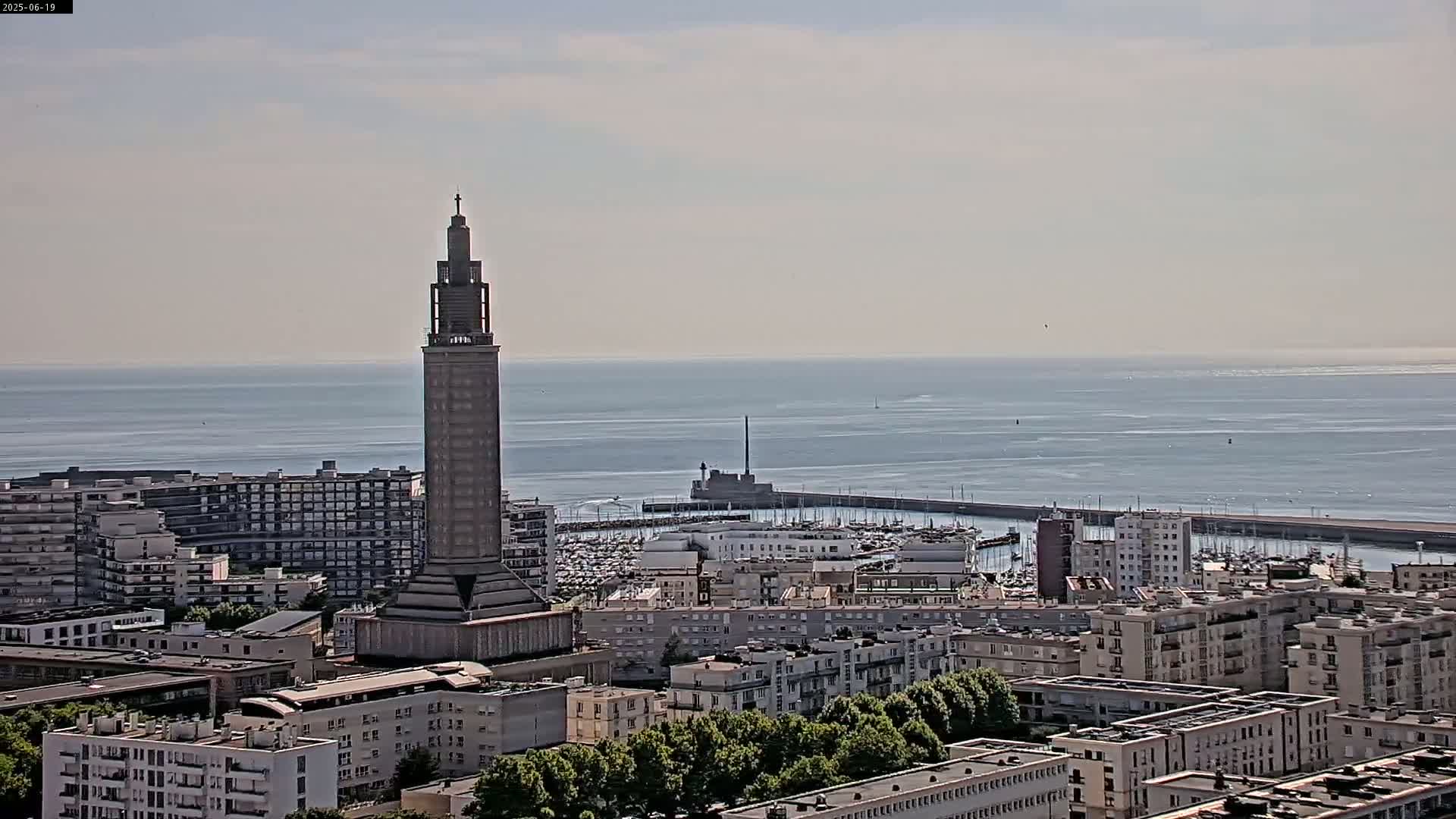 A tall, slender tower stands prominently in a cityscape overlooking a calm ocean on a hazy day.