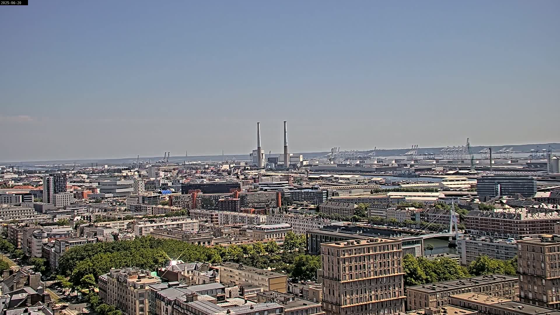 A sprawling cityscape under a clear sky shows a mix of residential and industrial buildings, including two prominent smokestacks and a large port area in the distance.
