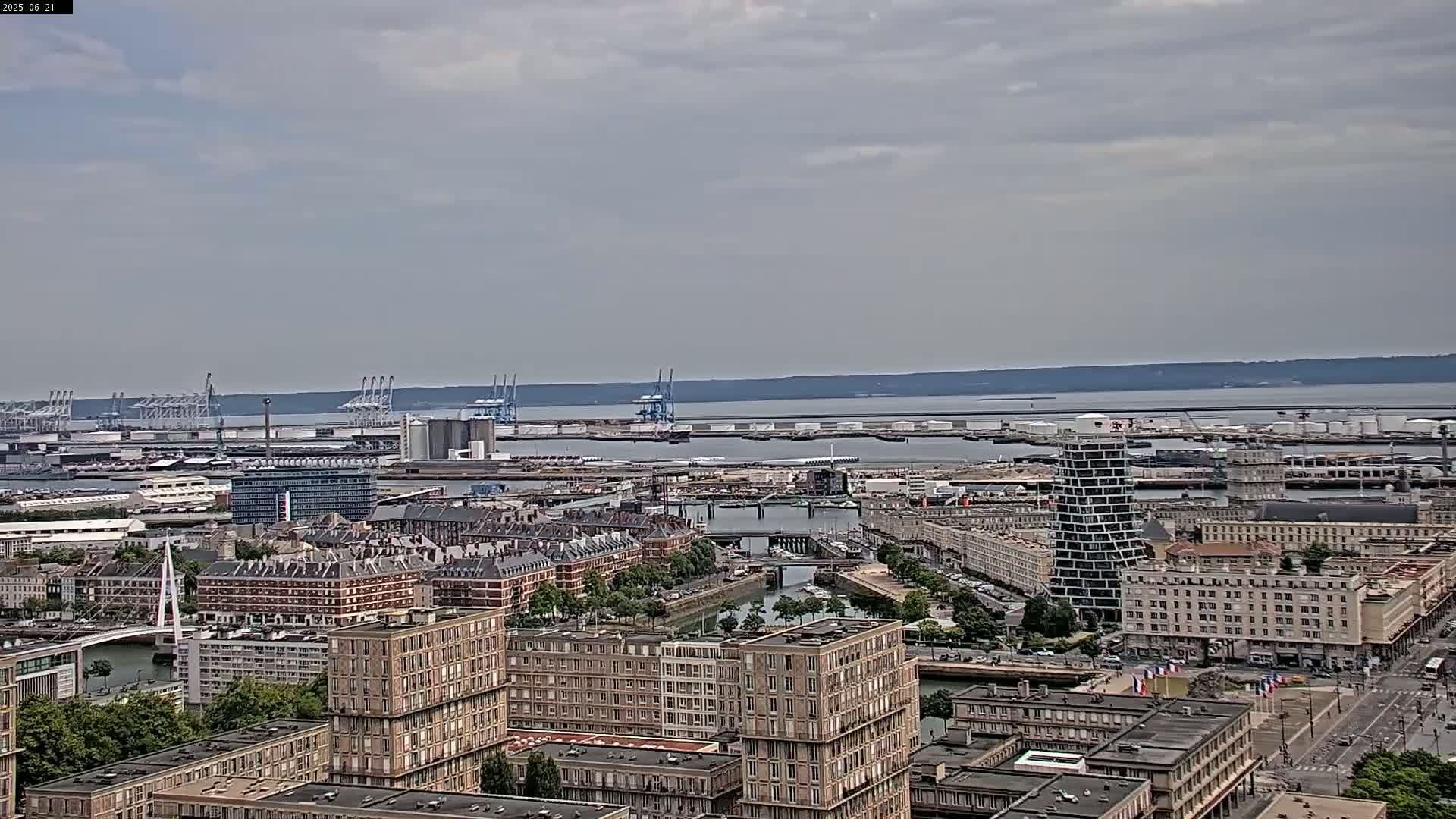 A city harbor with numerous buildings, cranes, and docked ships is visible under an overcast sky.