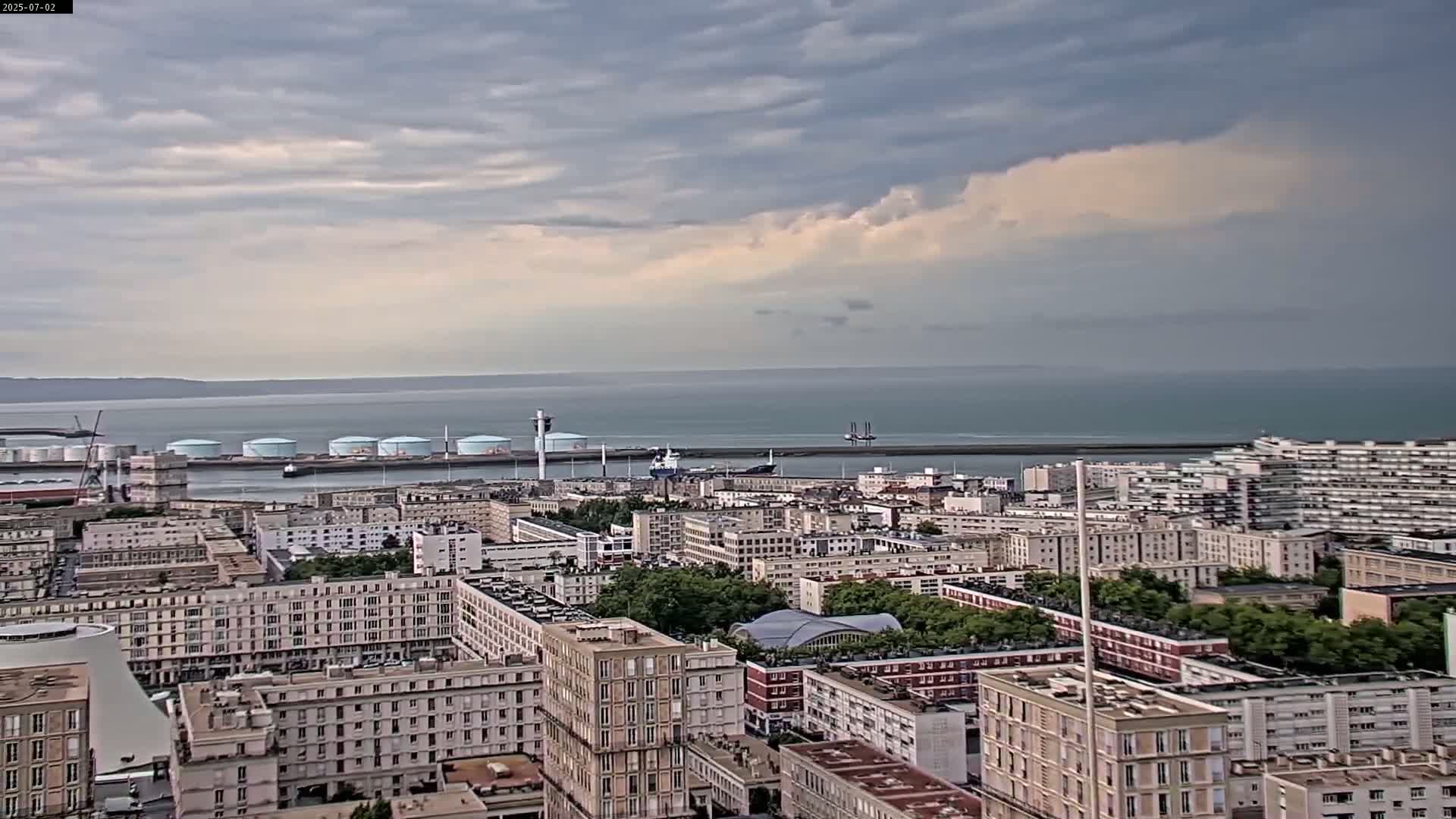 A coastal city with many multi-story buildings is shown under a mostly cloudy sky with a partially sunny horizon.