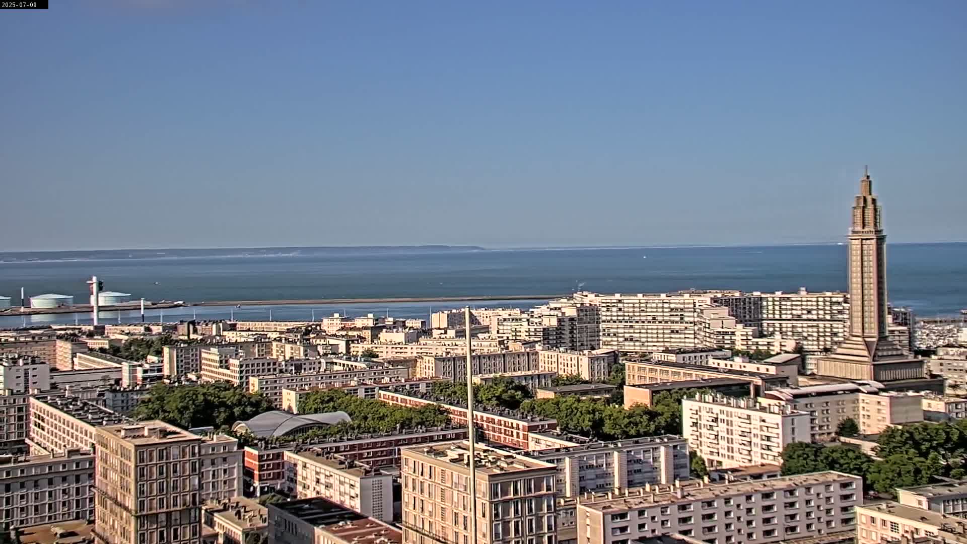 A coastal city with many apartment buildings, a tall tower, and a port is visible on a clear, sunny day.