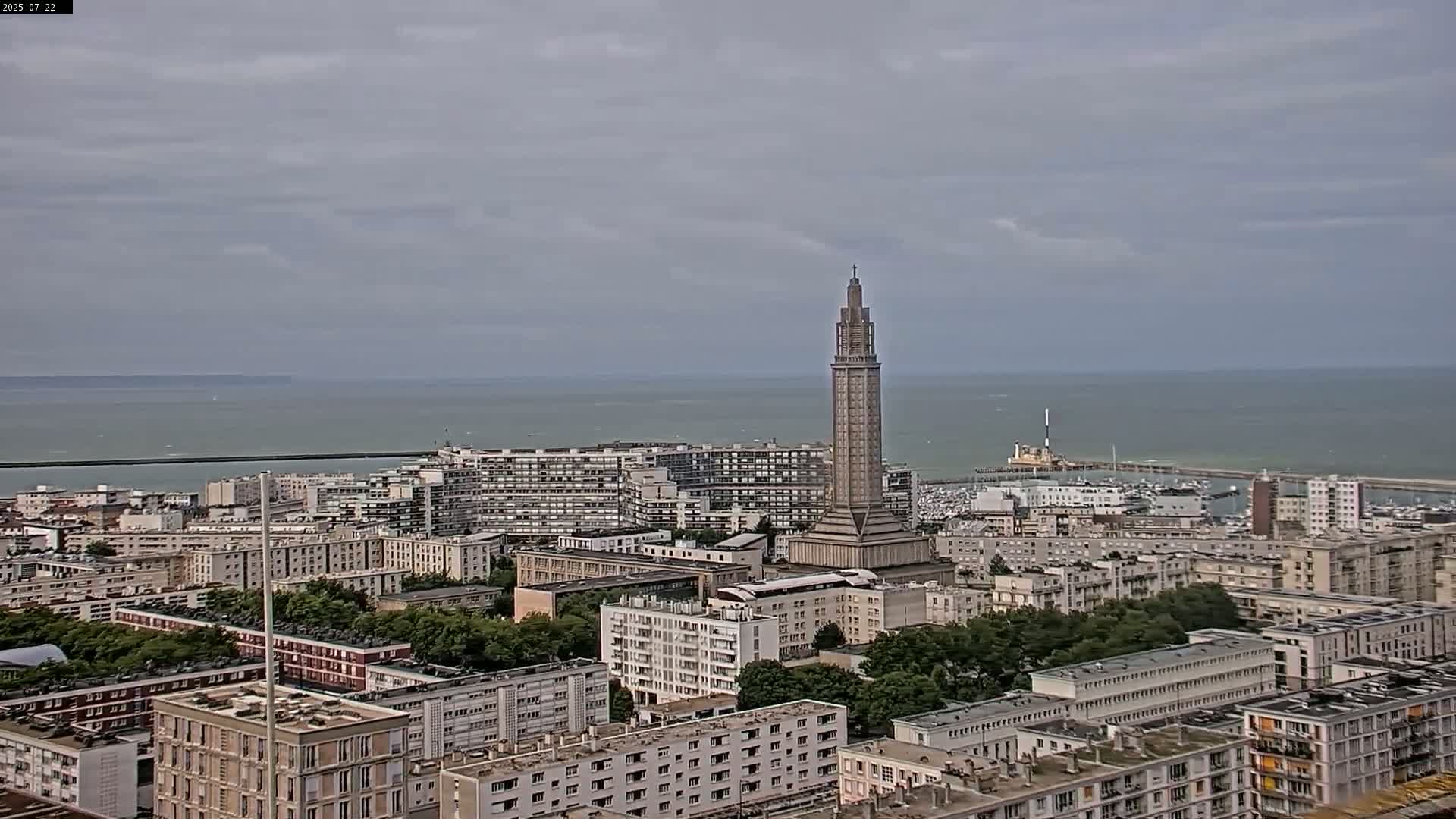A tall, slender tower stands in the midst of a city overlooking a calm, gray-sky ocean.