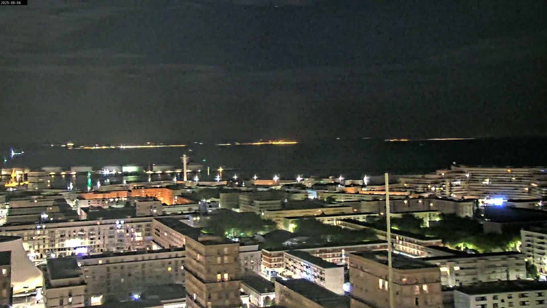 A nighttime aerial view shows a city's illuminated buildings and port area next to a dark body of water with faintly visible lights across the horizon under a mostly clear sky.