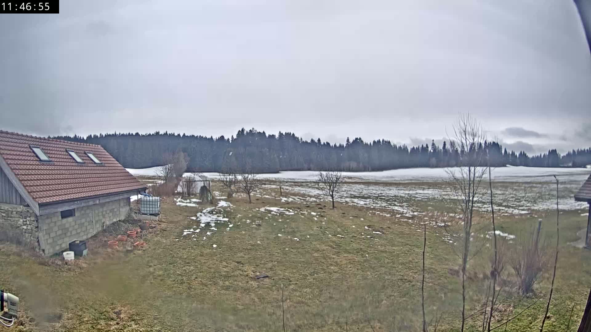 A stone and brown-roofed building sits in a field with patchy snow, overlooking a line of trees under an overcast sky.