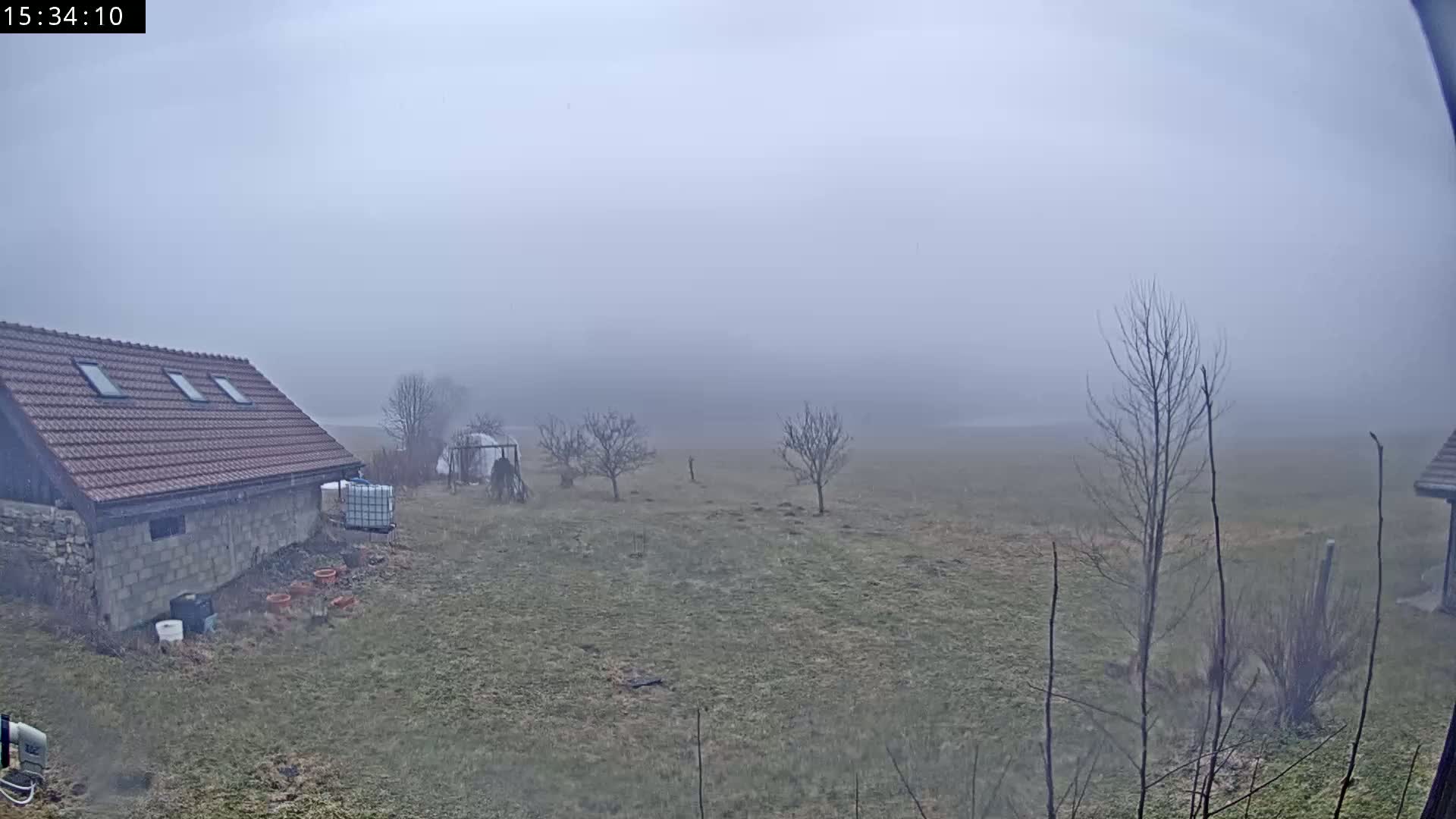 A stone building with a red tile roof sits in a foggy field with a few bare trees and some gardening supplies.