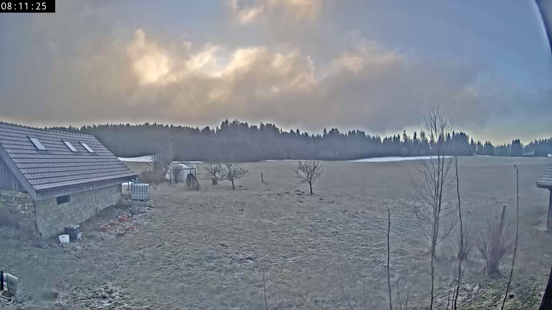 A stone building with a tiled roof sits beside a frost-covered field, backed by a line of dark trees under a partly cloudy sky.