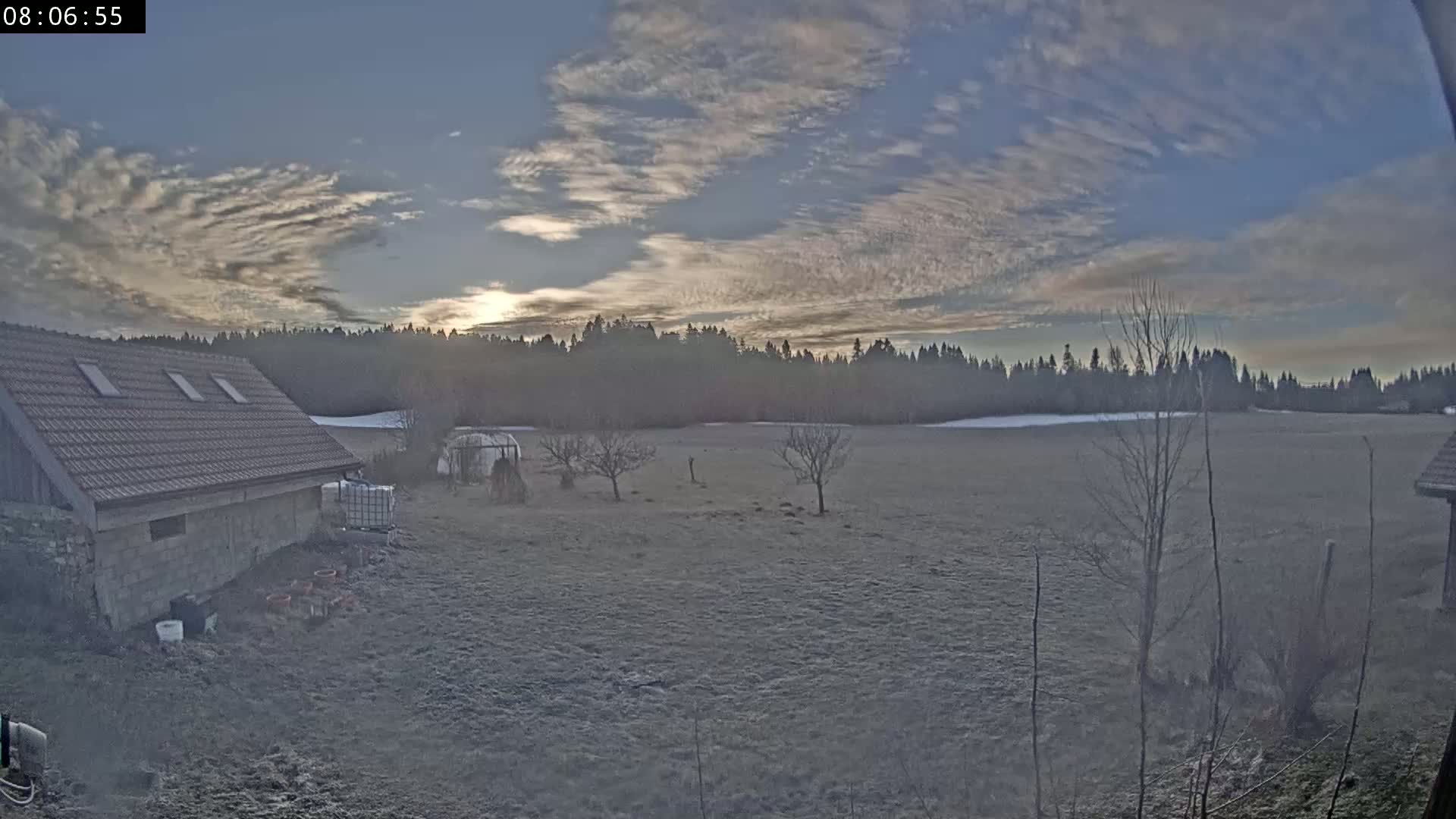 A stone building with a tiled roof sits beside a frosty field, under a partly cloudy, sunrise sky.