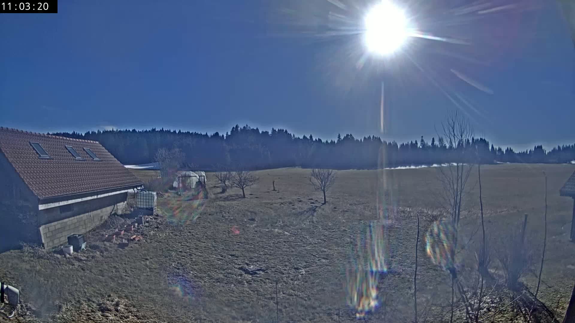 A red-roofed building sits beside a mostly barren field under a bright, sunny sky, with a line of dark trees in the distance.