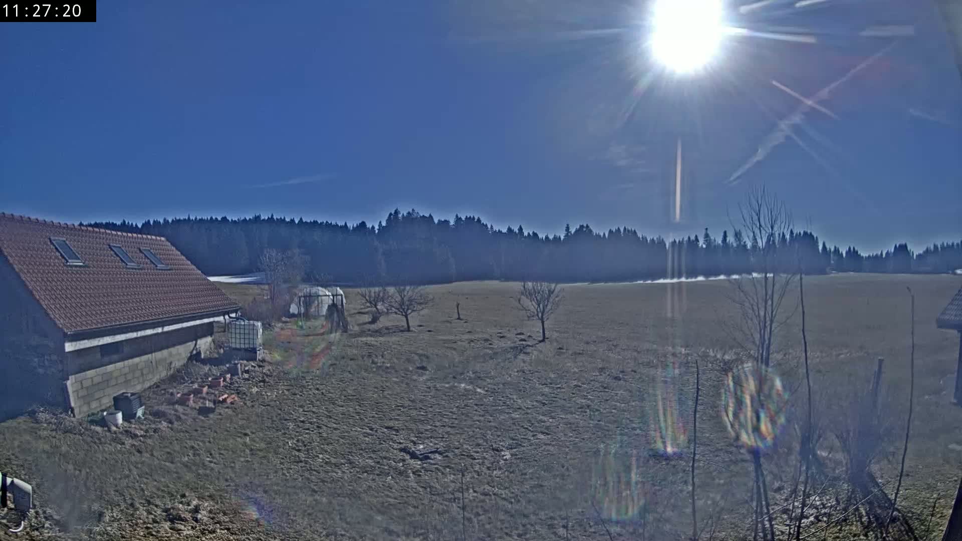 A brown-roofed building sits near a bare, brown field on a sunny day, with a line of dark trees in the background.