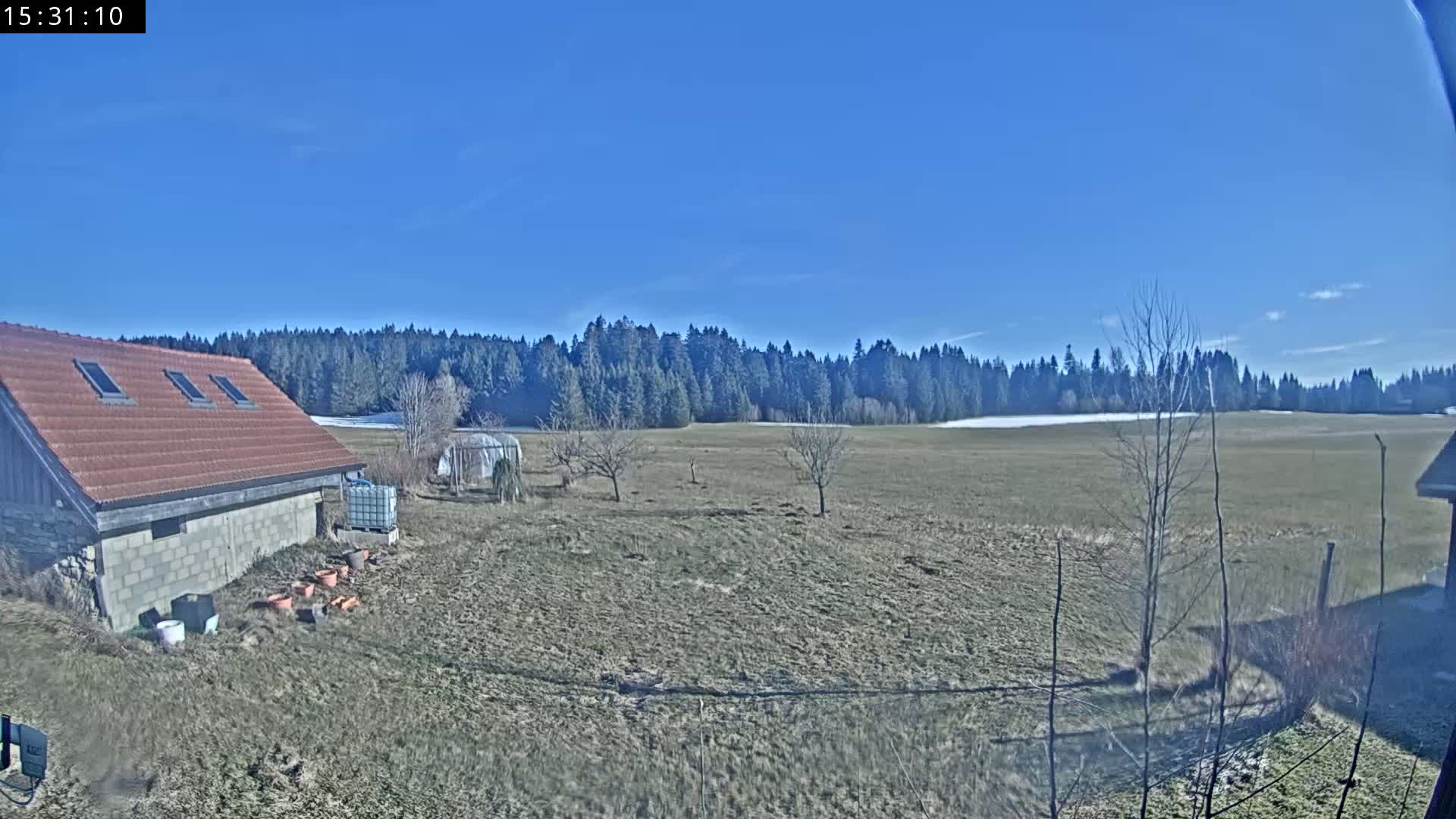 A brown-roofed building sits beside a mostly brown field, with a line of dark green trees in the background under a clear blue sky.