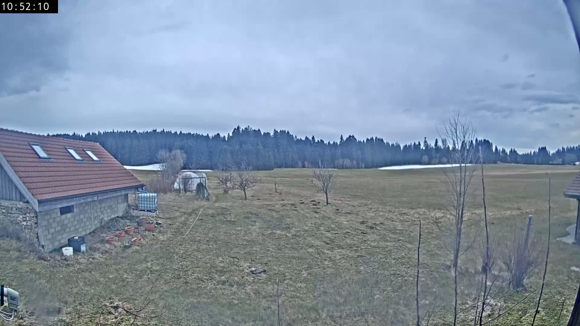 A stone building with a red tile roof sits in a field under an overcast sky, with a line of trees in the background.