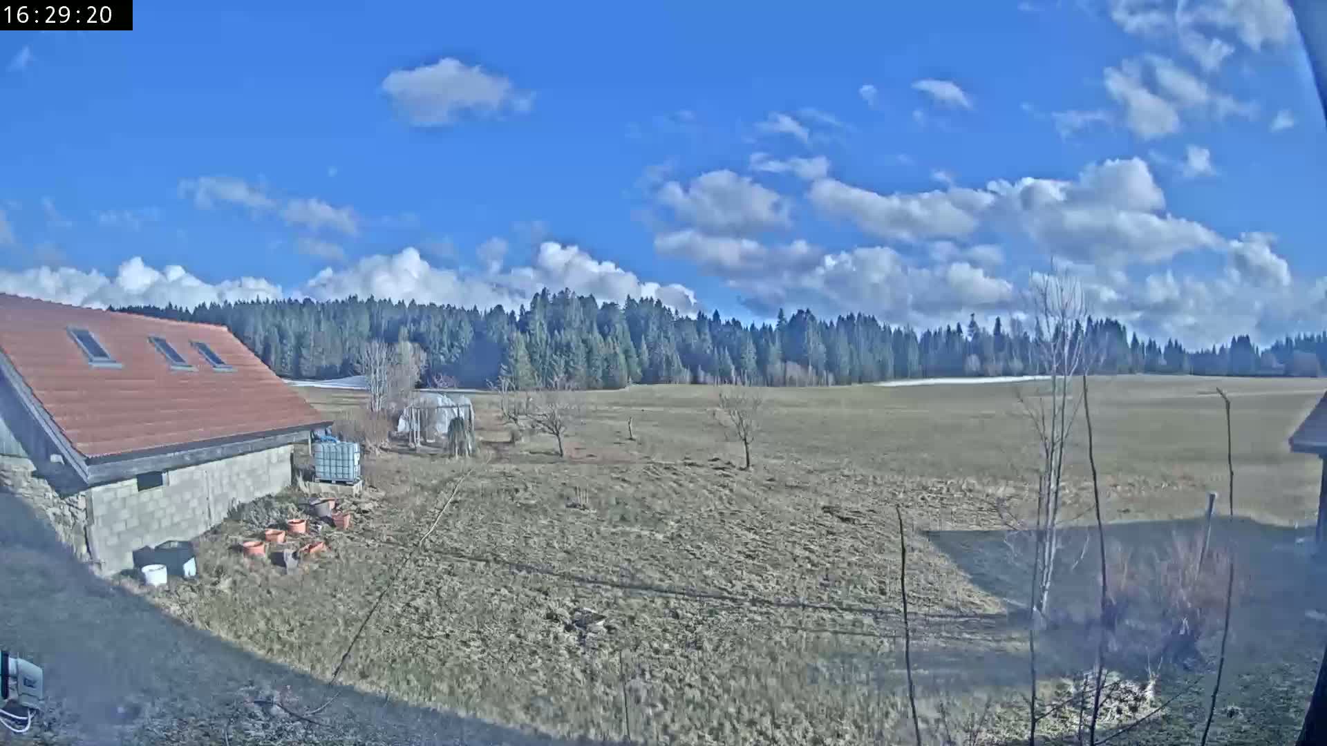A partly cloudy blue sky overlooks a rural scene of a brown-roofed building, a plowed field, and a line of evergreen trees in the distance.