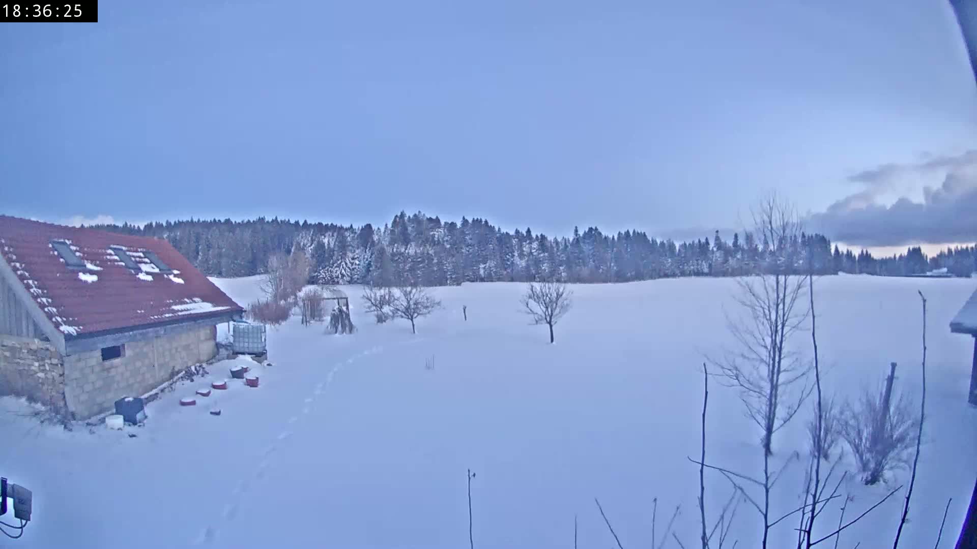 A snow-covered field with a stone building and leafless trees under a pale blue sky with some clouds.