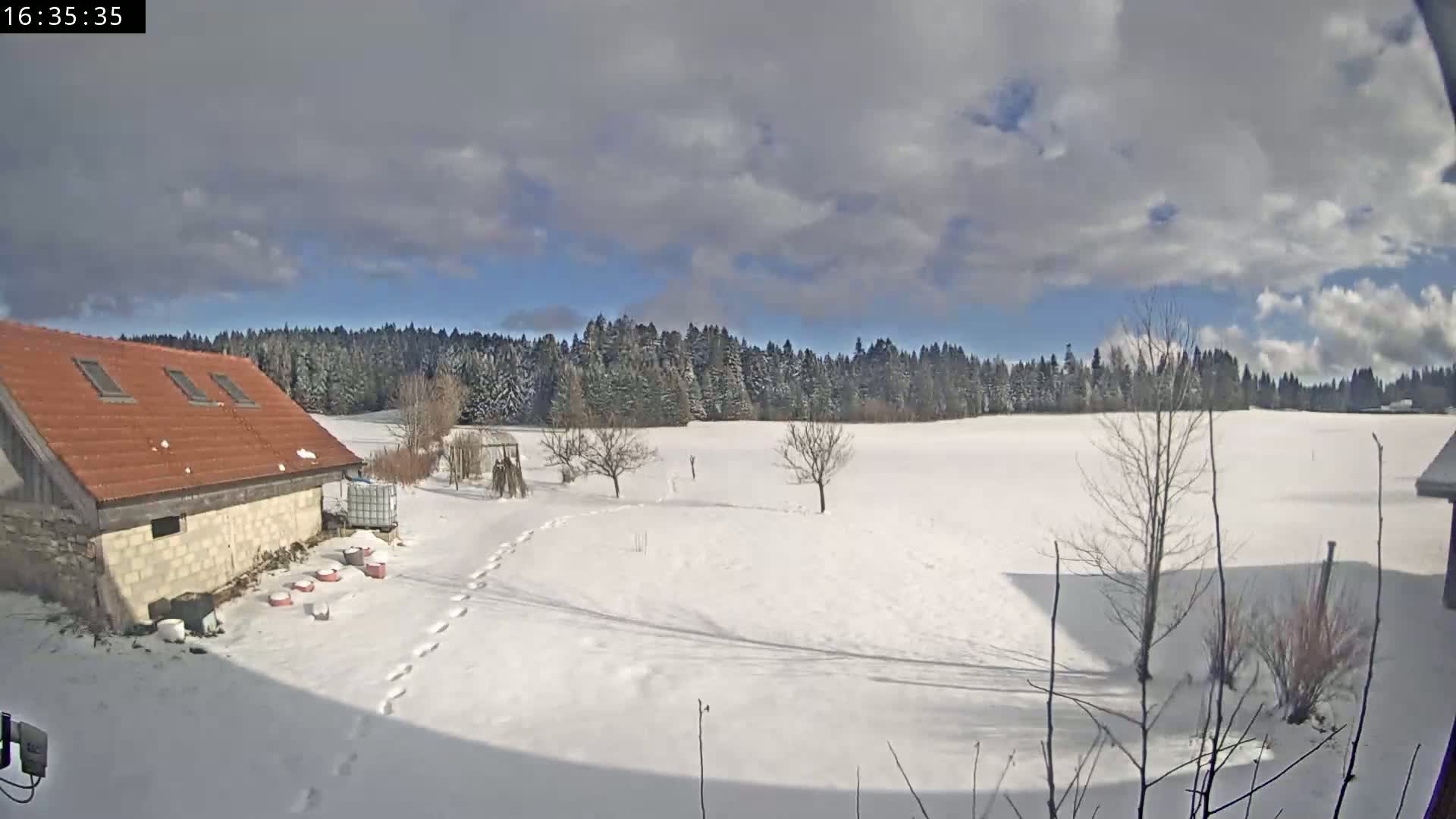 A snow-covered field with a few bare trees and a stone building with a red roof is visible under a partly cloudy sky.
