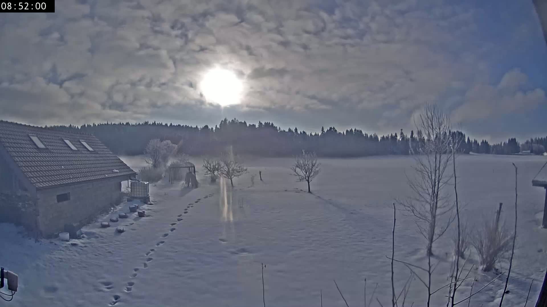 A stone building with a snow-covered yard and a sunlit, cloudy sky stretches beyond a snow-covered field and distant forest.