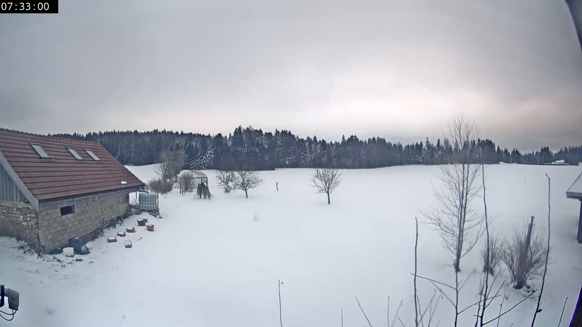 A snow-covered field stretches out before a stone building with a red tile roof, under an overcast sky, with a line of dark trees in the background.
