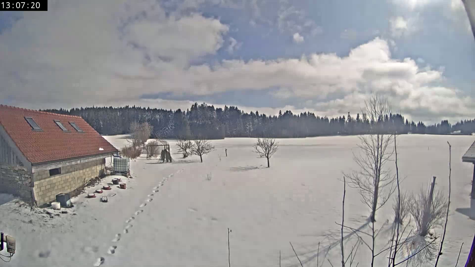 A snow-covered field with a few bare trees and a stone building with a red roof is visible under a partly cloudy sky.