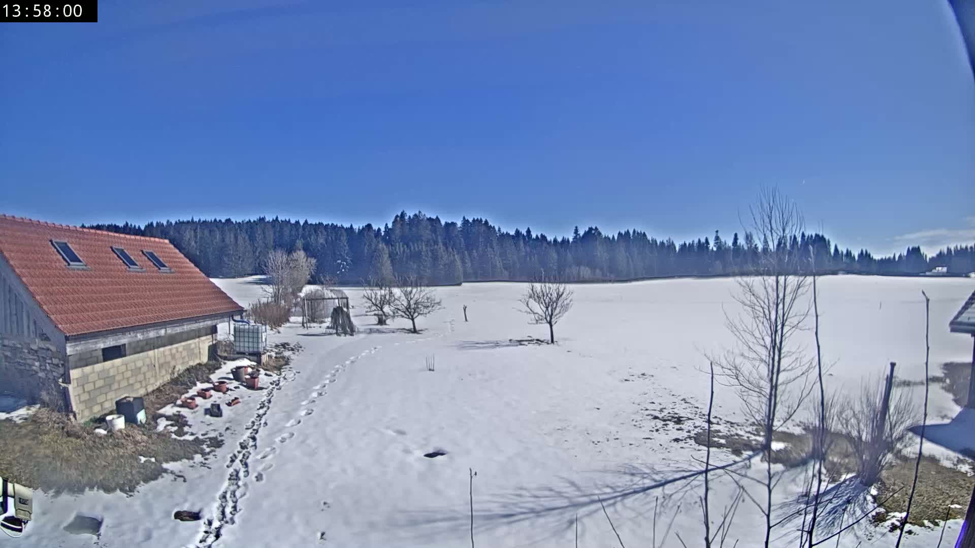 A snow-covered field stretches out before a low, red-roofed building, with a line of dark trees in the background under a clear blue sky.