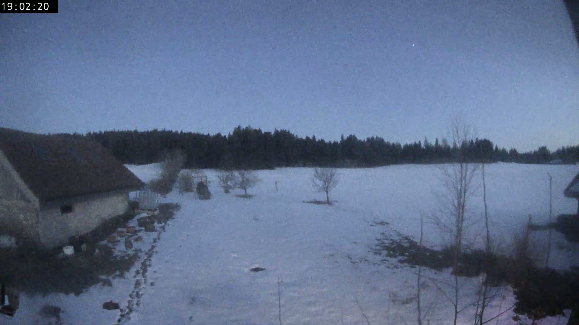 A snow-covered field stretches out before a low building under a twilight sky, bordered by a dark line of trees.