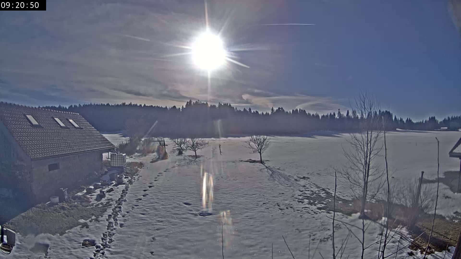 A snow-covered field with a building and some trees is bathed in bright sunlight under a mostly clear sky.