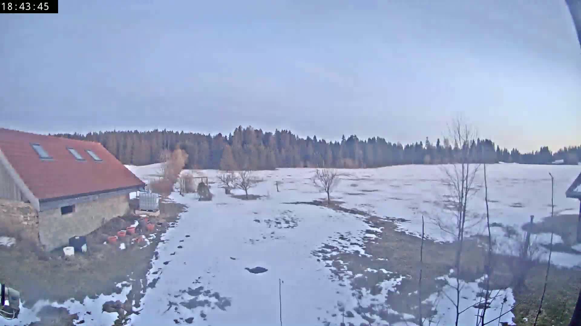 A stone building with a red roof sits near a snow-covered field, with a line of trees in the background under a clear, pale sky.