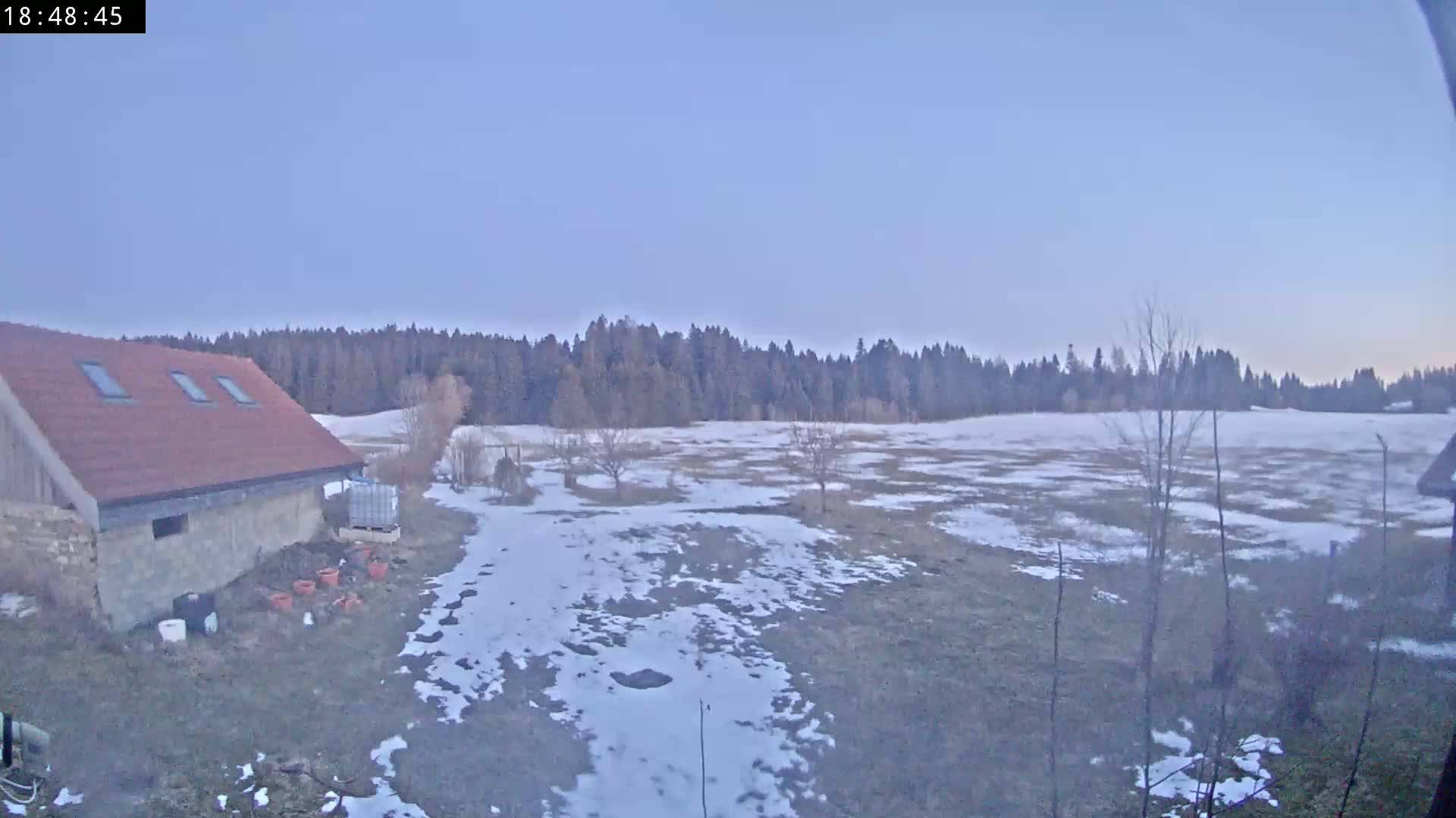 A stone building with a red roof sits in a snowy field, overlooking a line of dark trees under a pale, twilight sky.