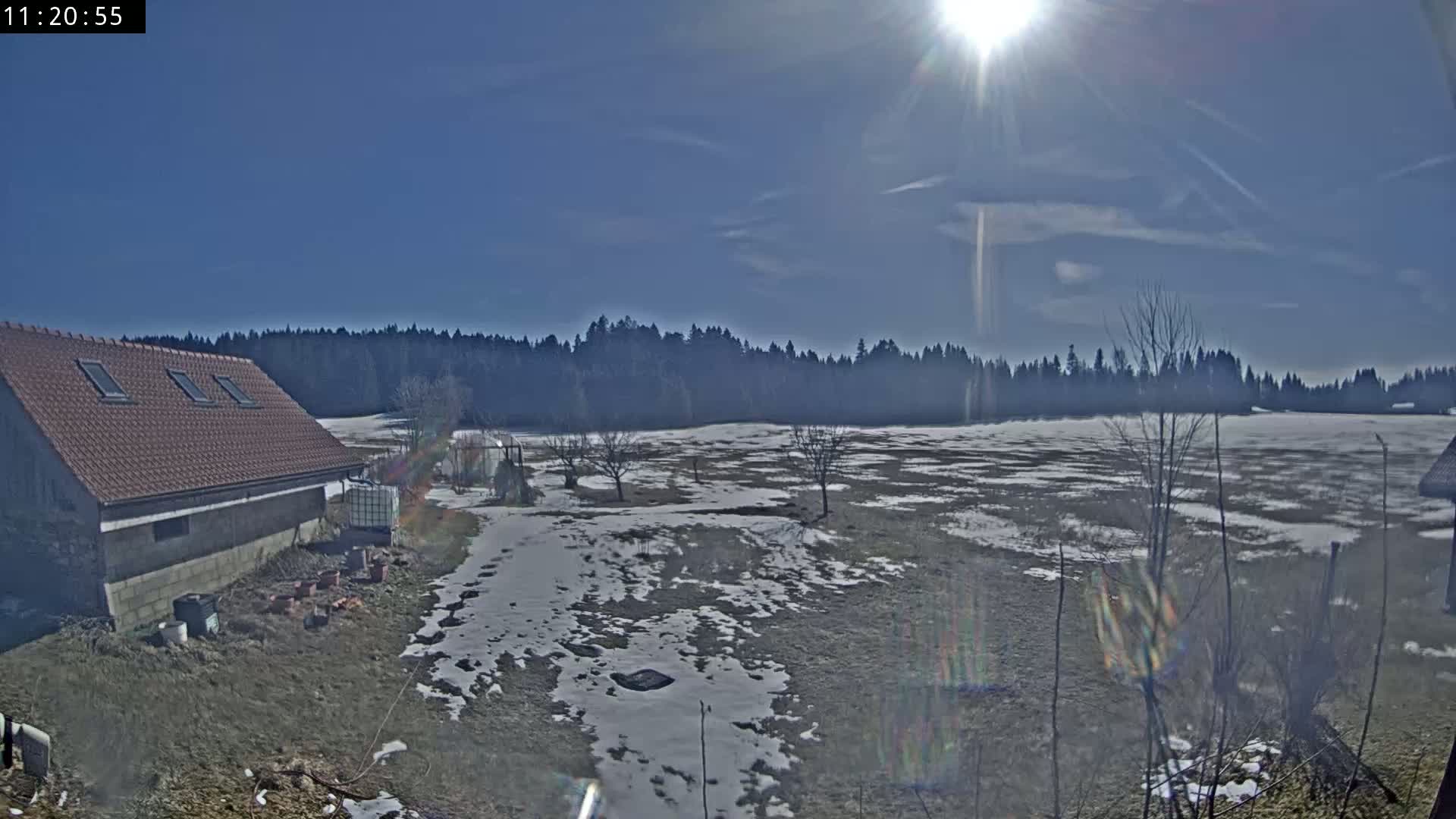 A partly sunny day reveals a snow-dusted field, a brown-roofed building, and a dark line of trees in the background.