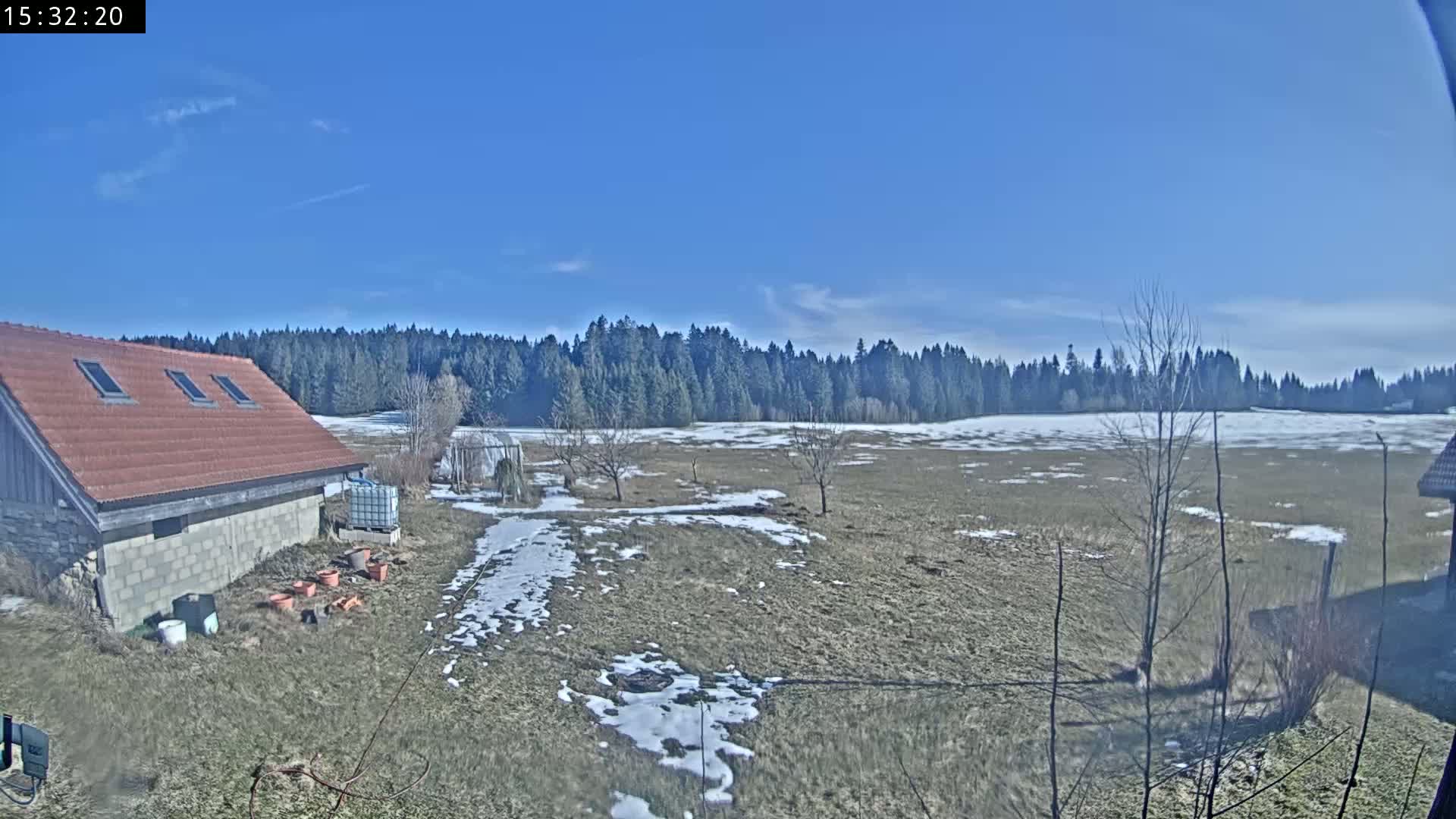 A mostly clear blue sky looks over a flat, snow-dusted field, a small building with a reddish-brown roof, and a line of evergreen trees.