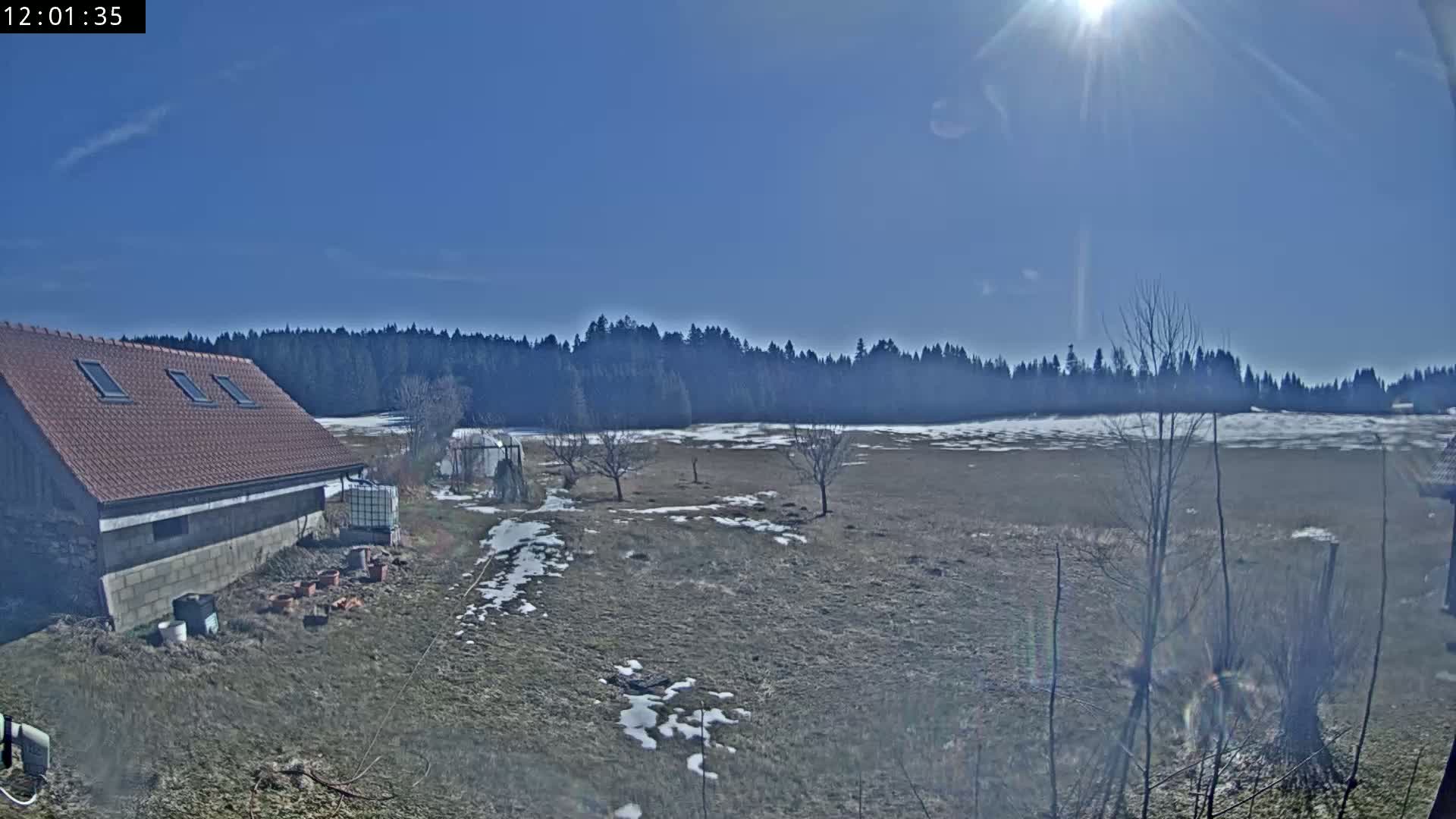A brown-roofed building sits near a snow-dusted field, bordered by a dark line of trees under a bright, sunny sky.