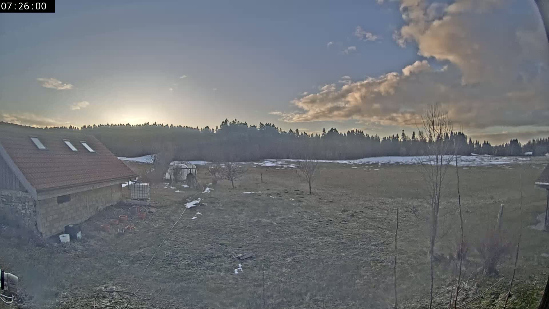 A brown-roofed building sits near a field with patches of snow, under a partly cloudy sky as the sun rises behind a line of trees.