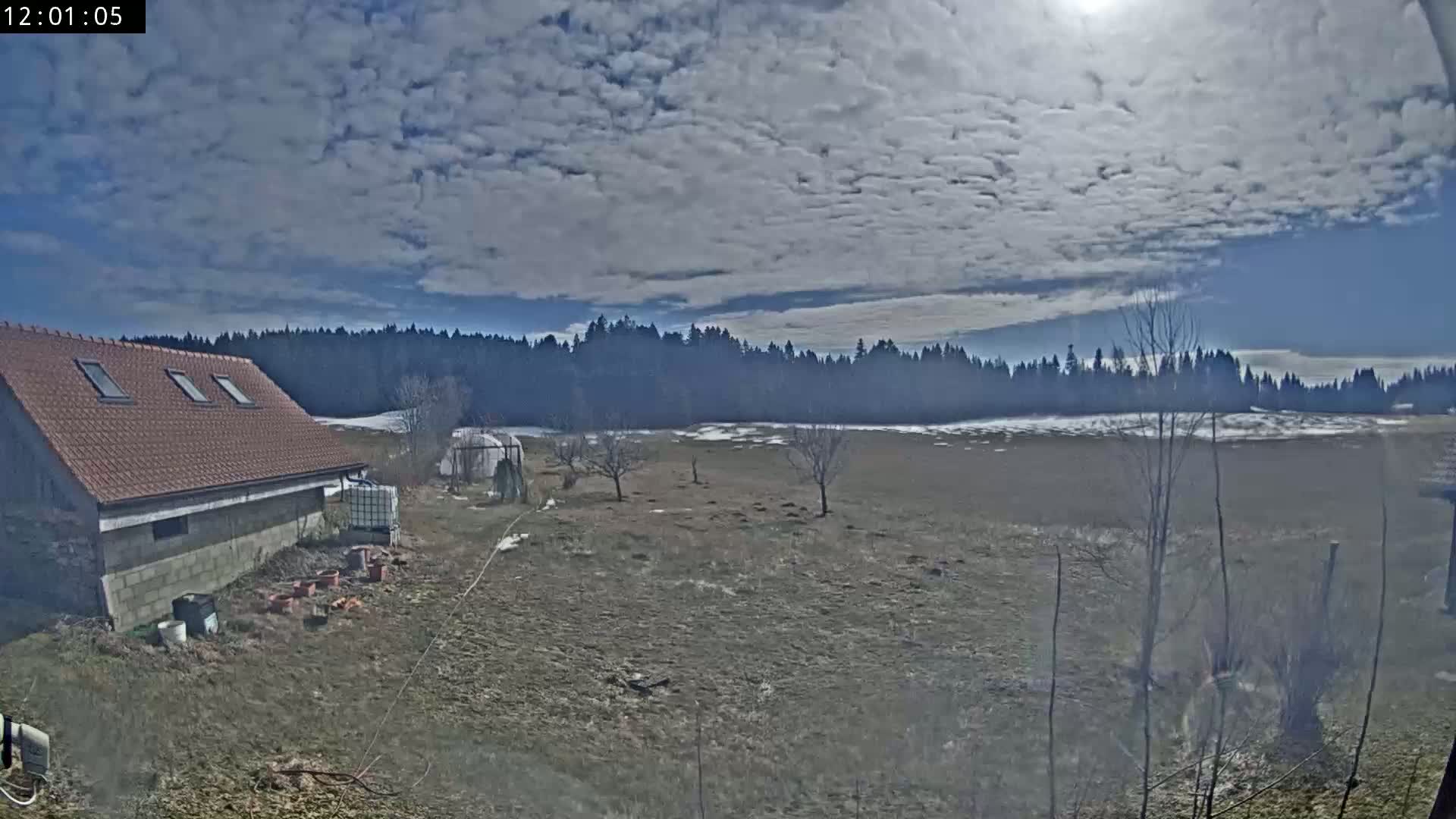 A brown-roofed building sits near a mostly barren field with a few small trees and patches of snow, under a partly cloudy sky.