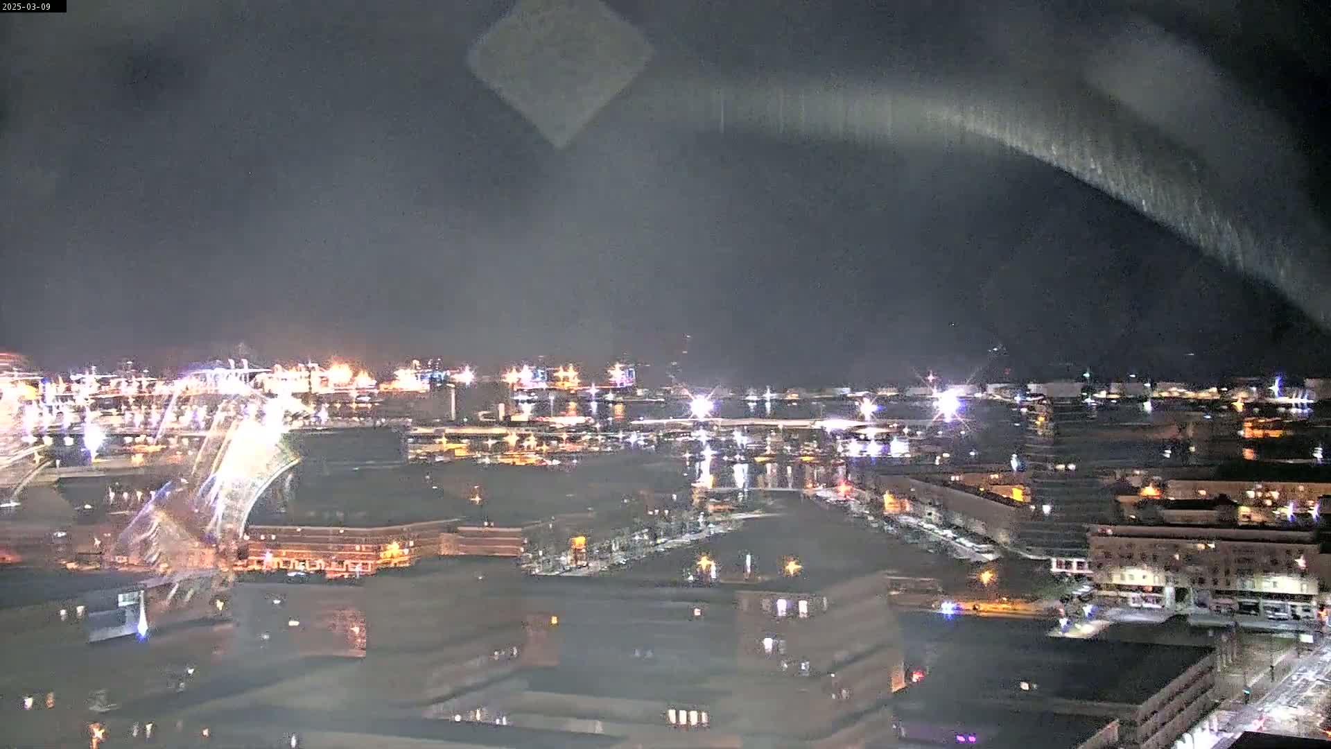 A nighttime view of a city harbor and waterfront, under a dark sky with faint streaks of light.