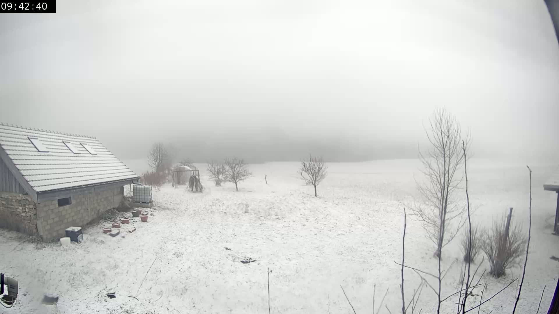 A snow-covered field stretches out behind a small stone building on a foggy day.