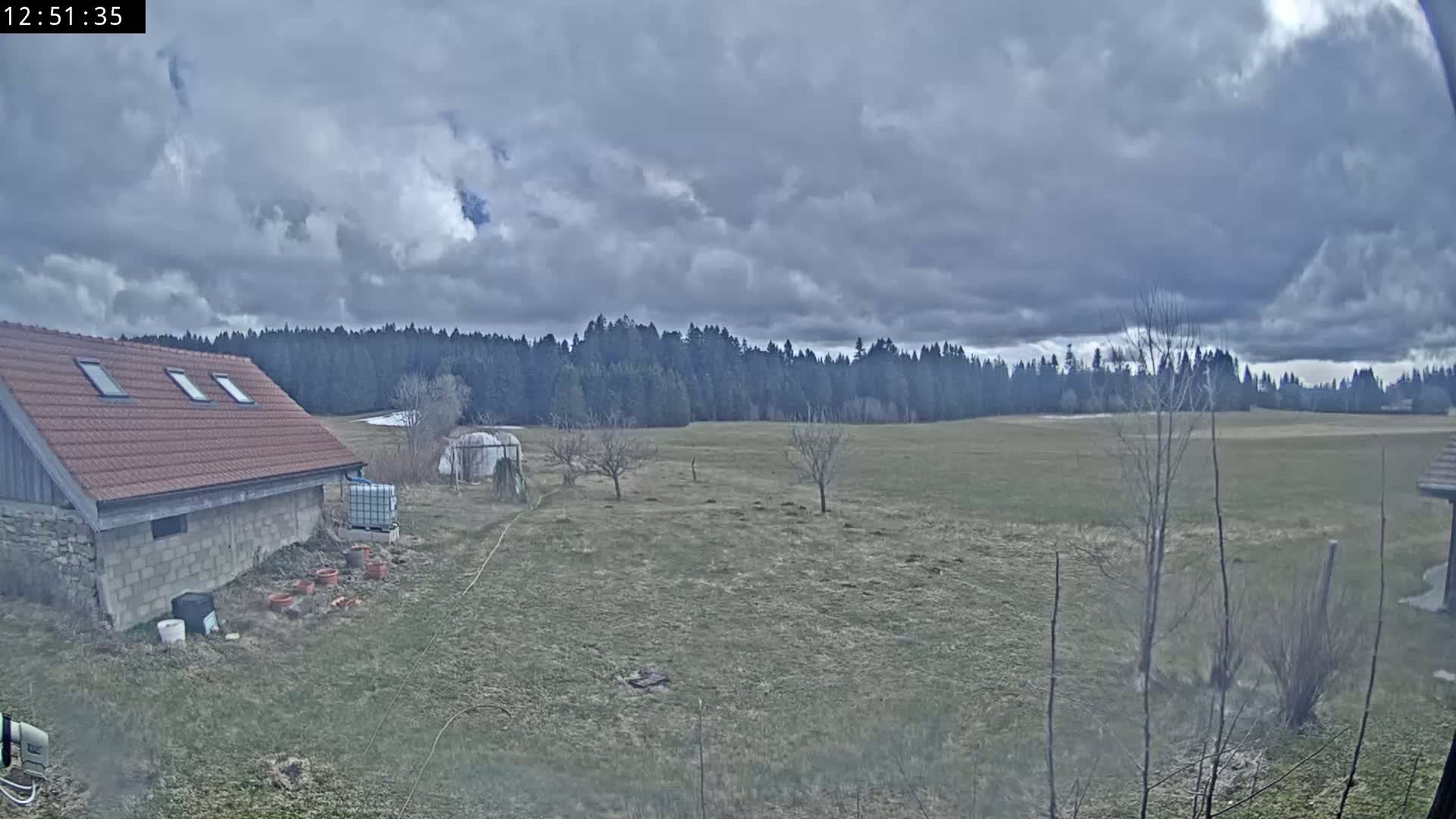 A brown-roofed building sits near a field, sparsely dotted with trees, under a cloudy sky.