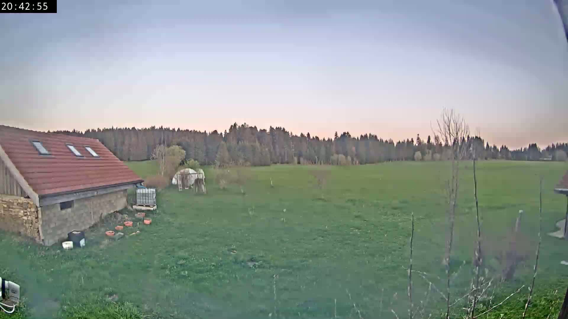 A red-roofed building sits on a grassy yard next to a large, green field bordered by a line of trees under a twilight sky.