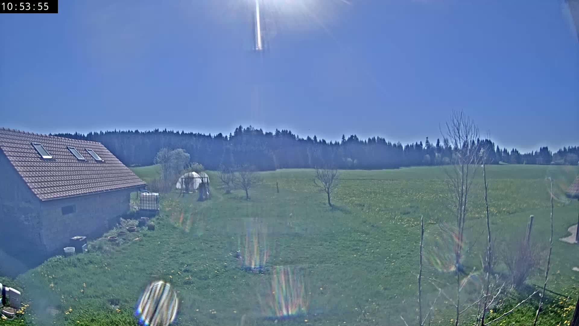 A stone building with a red tile roof sits on a grassy yard, overlooking a sunny field dotted with trees and a distant forest under a clear blue sky.
