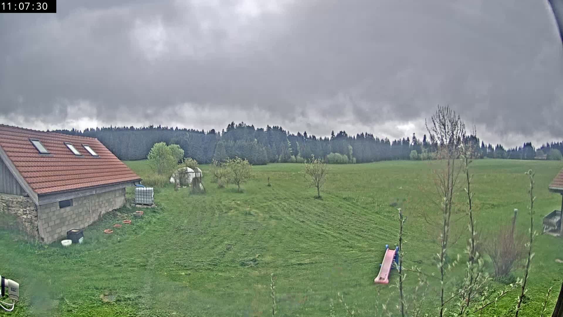 A red-roofed building sits in a grassy field under an overcast sky, with a line of trees in the distance and a small pink slide visible in the foreground.