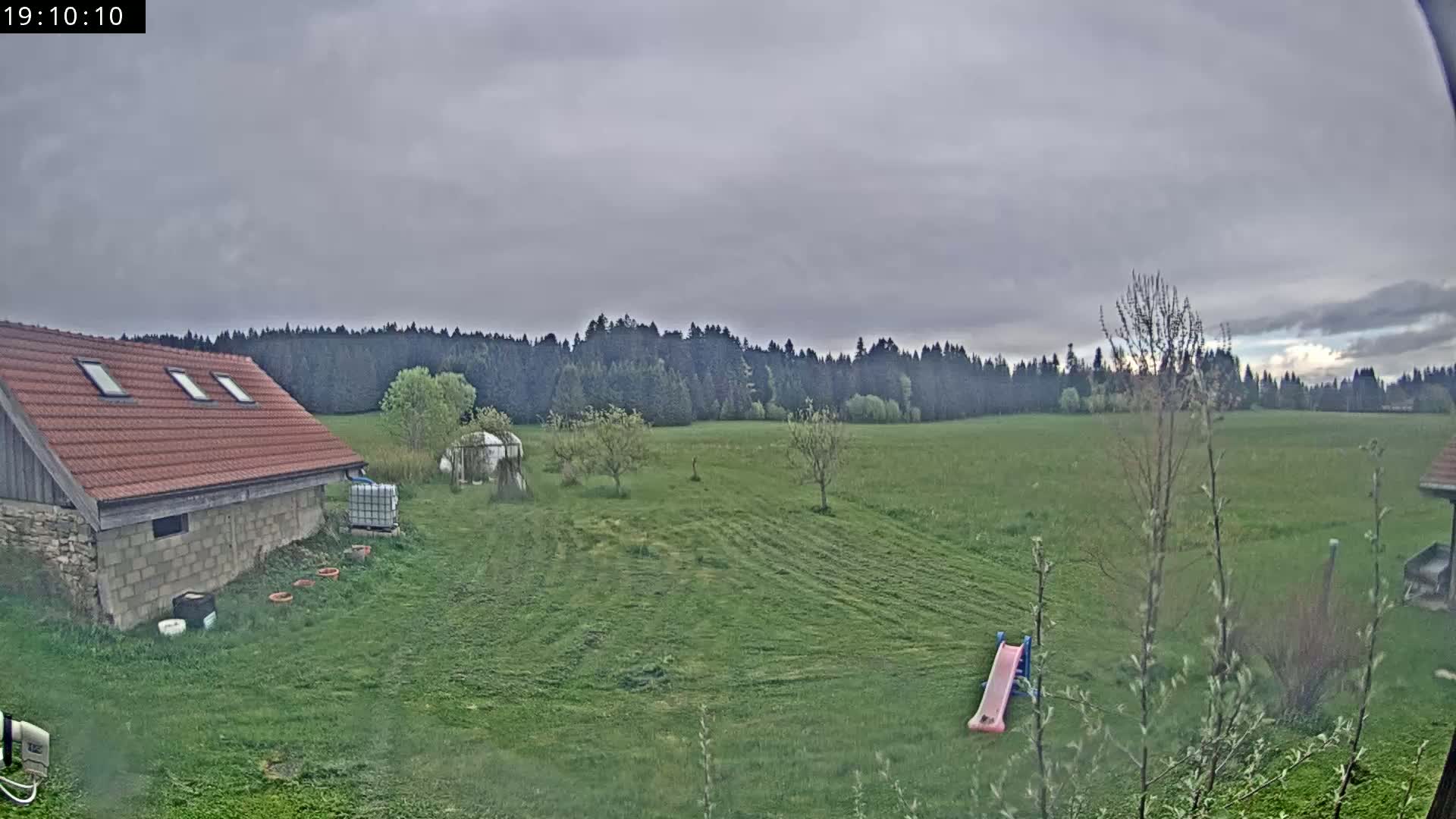 A red-roofed building sits in a grassy field under an overcast sky, with a line of trees in the distance and a small pink slide visible in the foreground.
