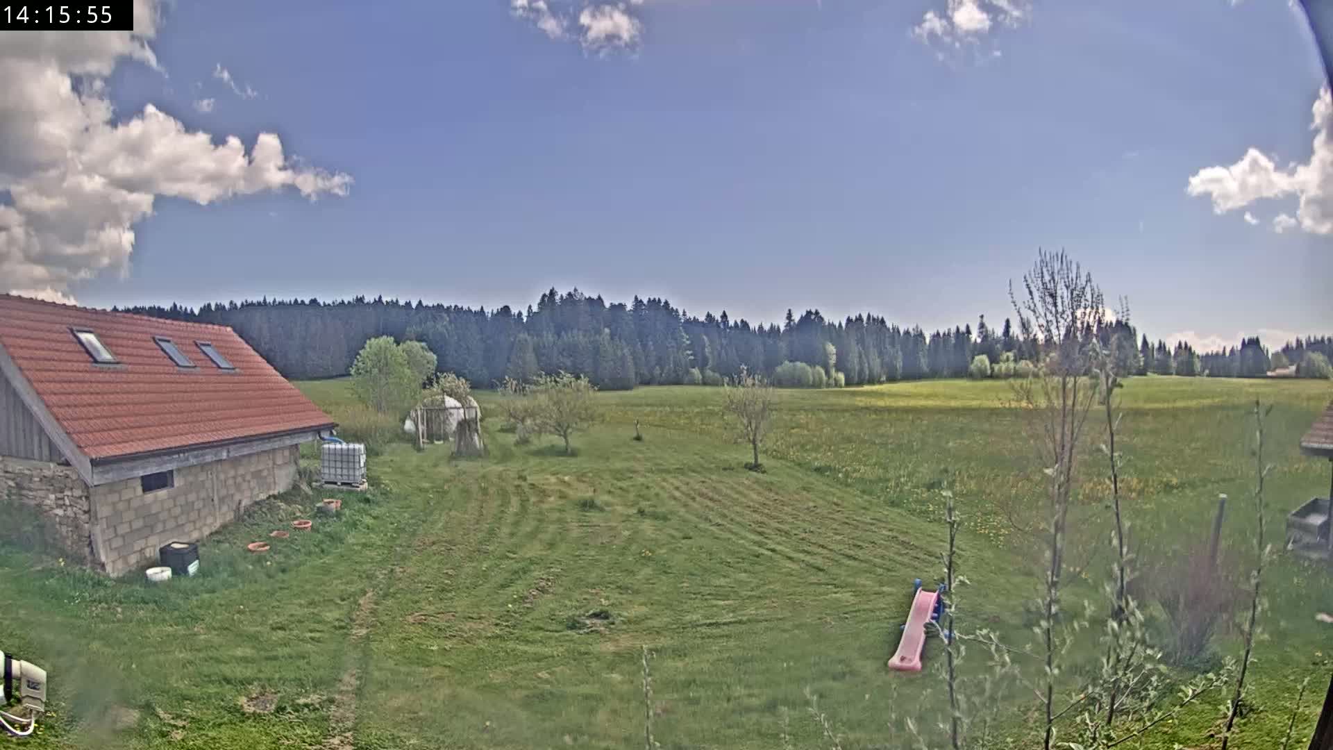 A red-roofed stone building sits on a grassy yard overlooking a large field and a distant forest under a partly cloudy blue sky.