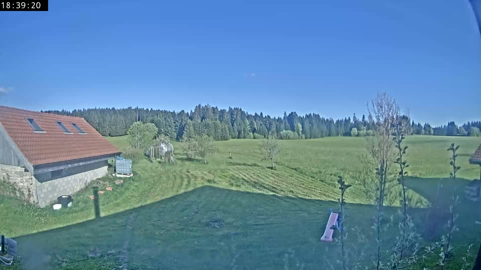 A red-roofed building sits near a grassy yard, overlooking a large, mown field bordered by a line of trees under a clear blue sky.