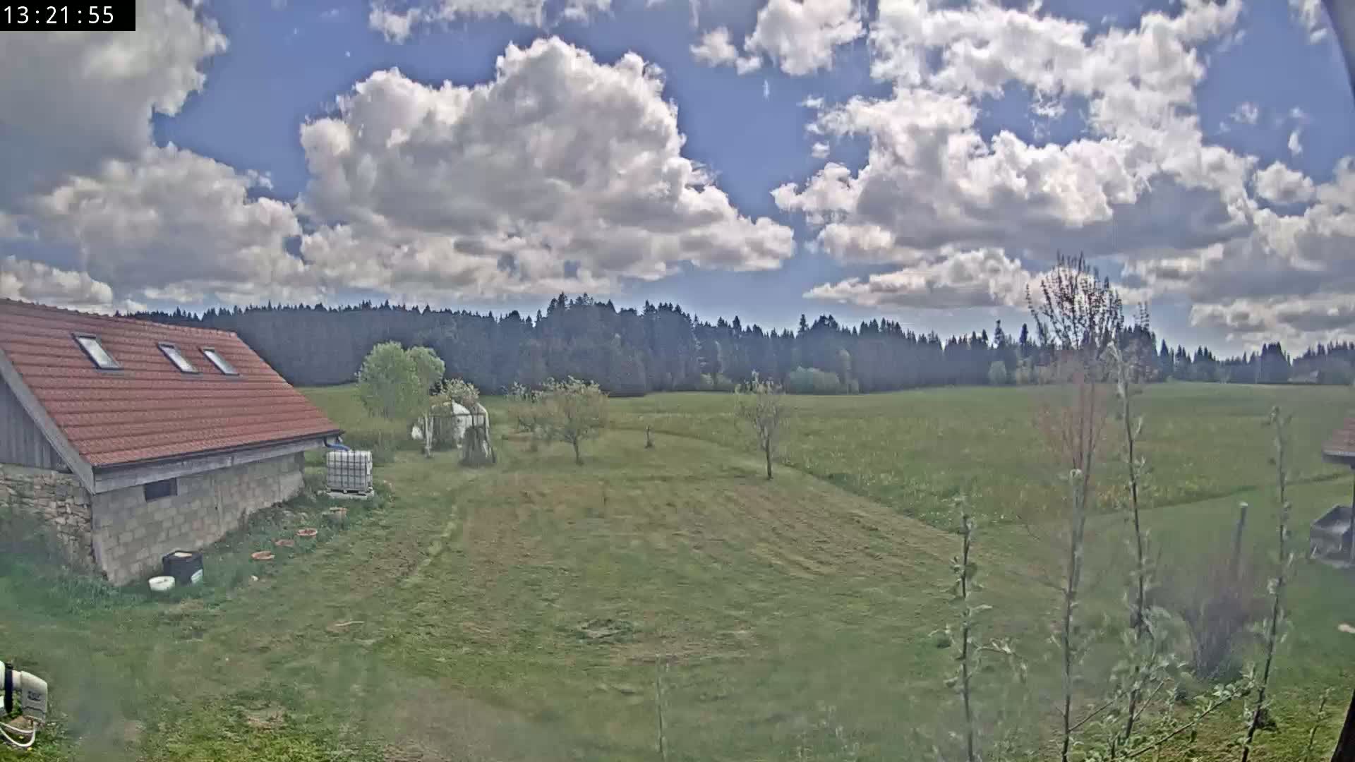 A red-roofed building sits in a grassy field under a partly cloudy sky, with a line of trees in the background.