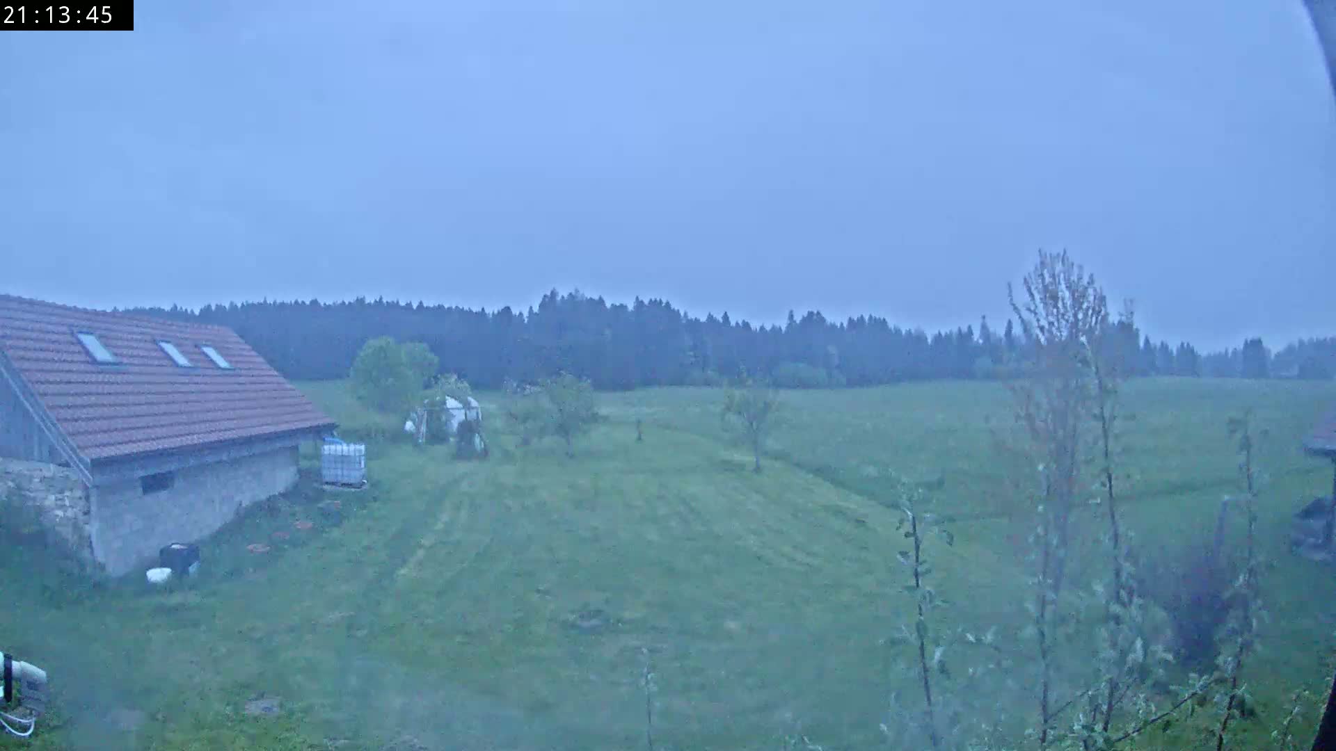 A low-lying building with a red tile roof sits on a grassy field, overlooked by a dark line of trees under an overcast sky.