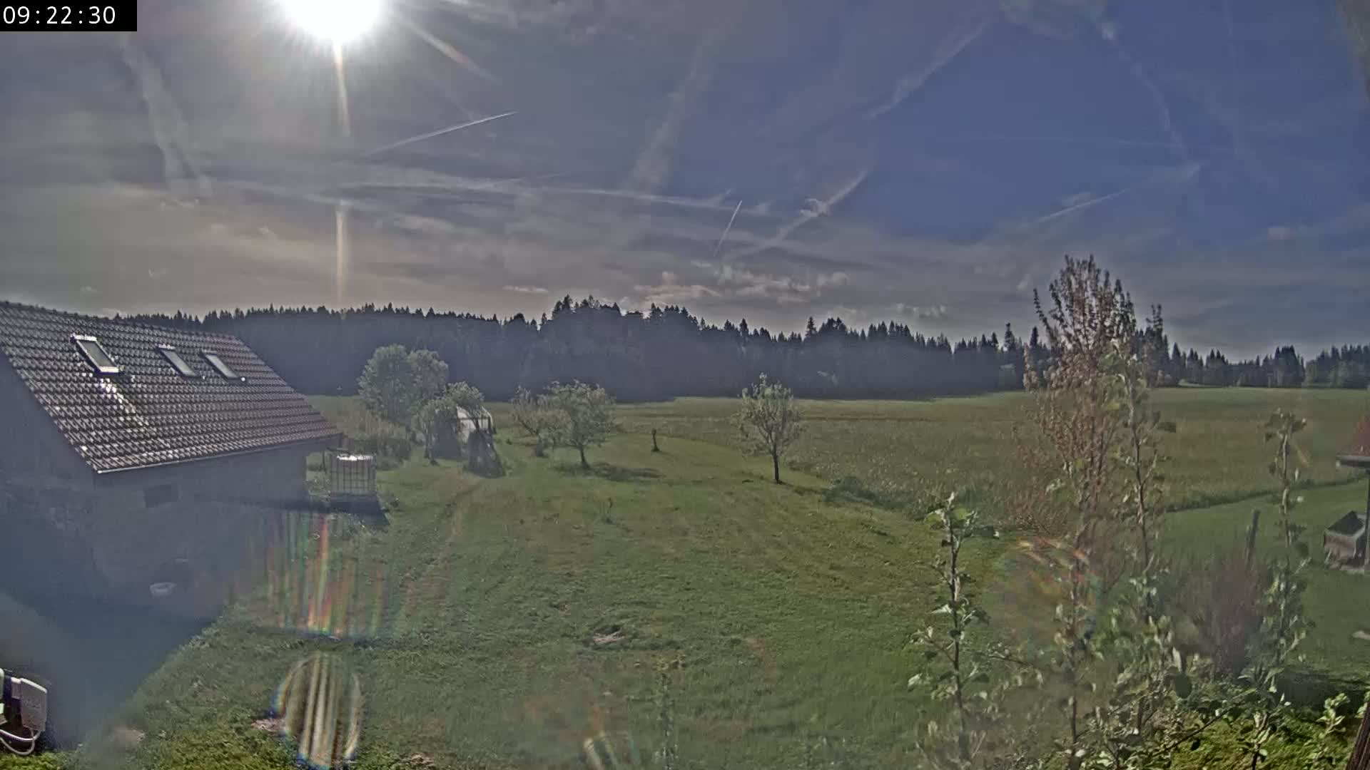A partly sunny day shows a view of a grassy field, a house with a tiled roof, and a line of trees in the background.