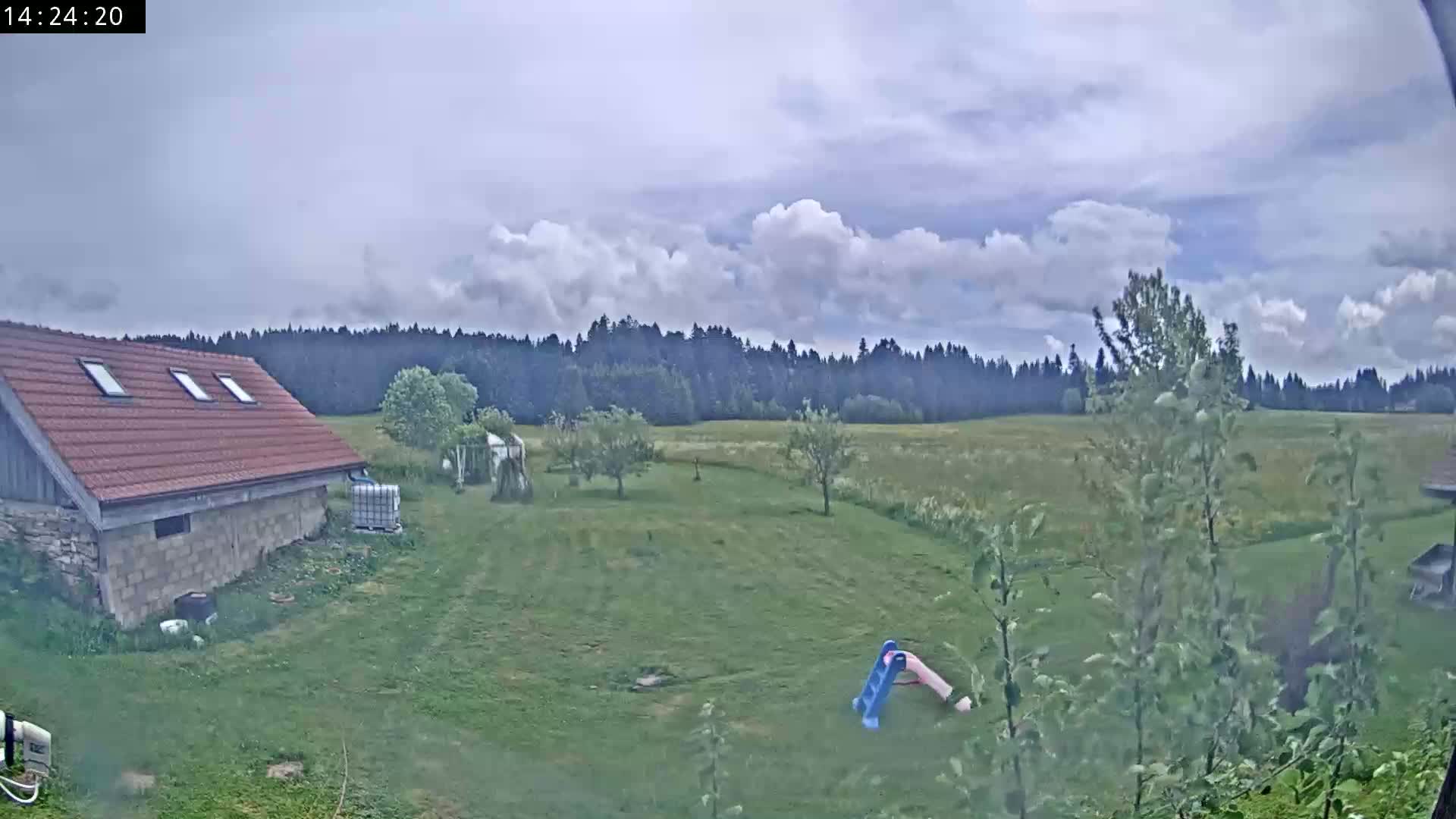 A partly cloudy day shows a red-roofed stone building, a grassy yard with a small slide, and a line of trees in the distance.