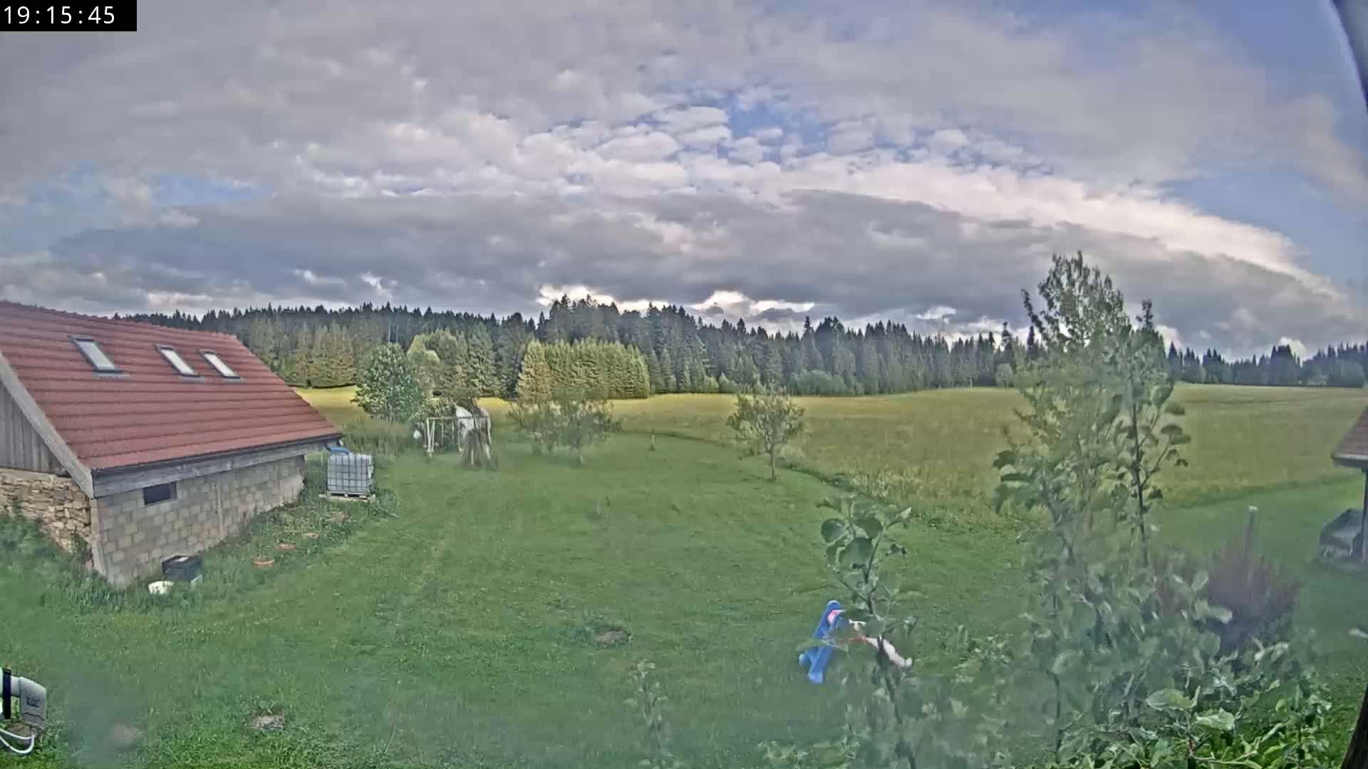 A red-roofed building sits on a grassy yard overlooking a field and a line of trees under a partly cloudy sky.