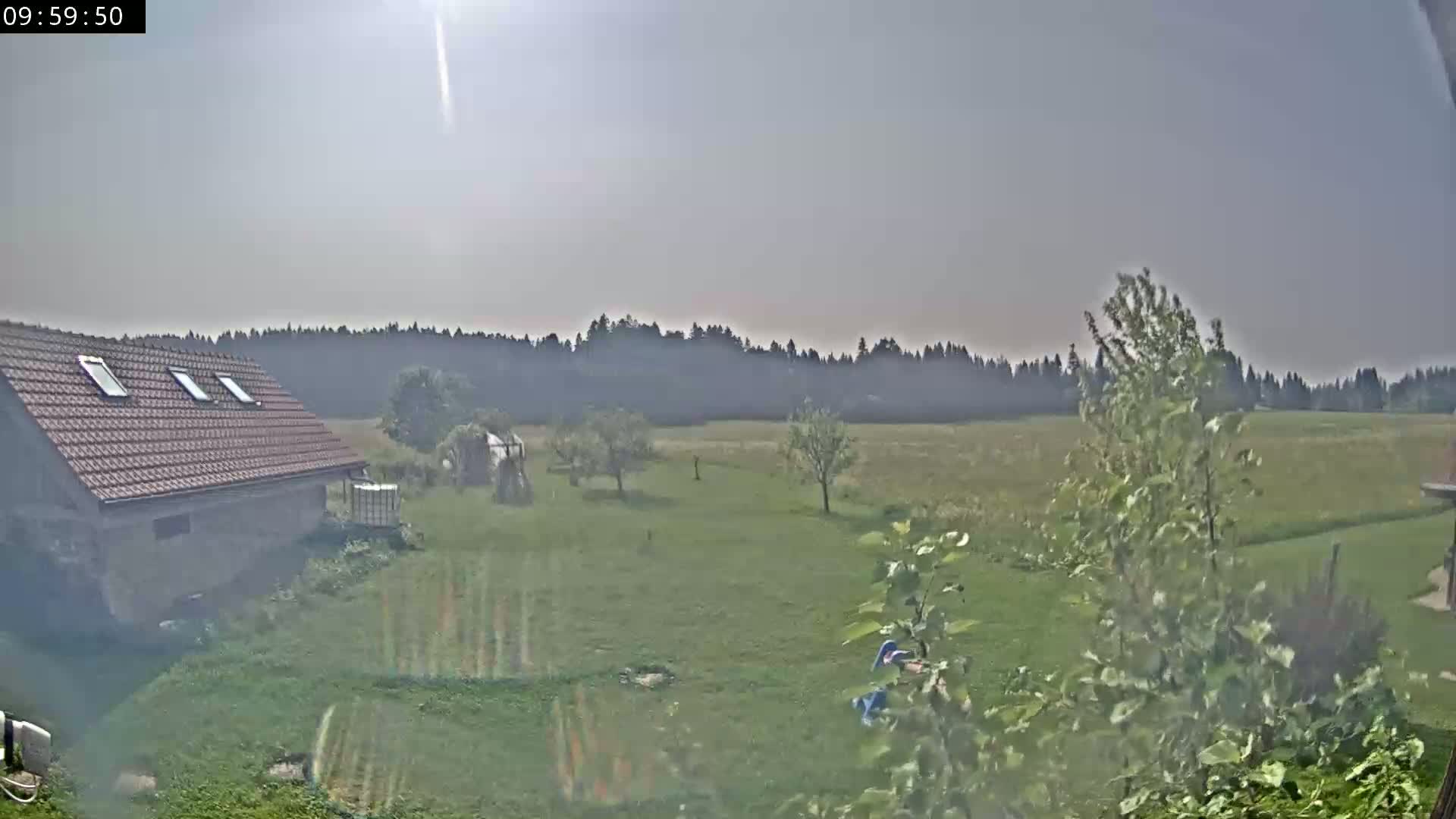 A hazy sunny day reveals a stone building with a brown roof, a grassy yard with some trees and a distant line of trees under a mostly clear sky.