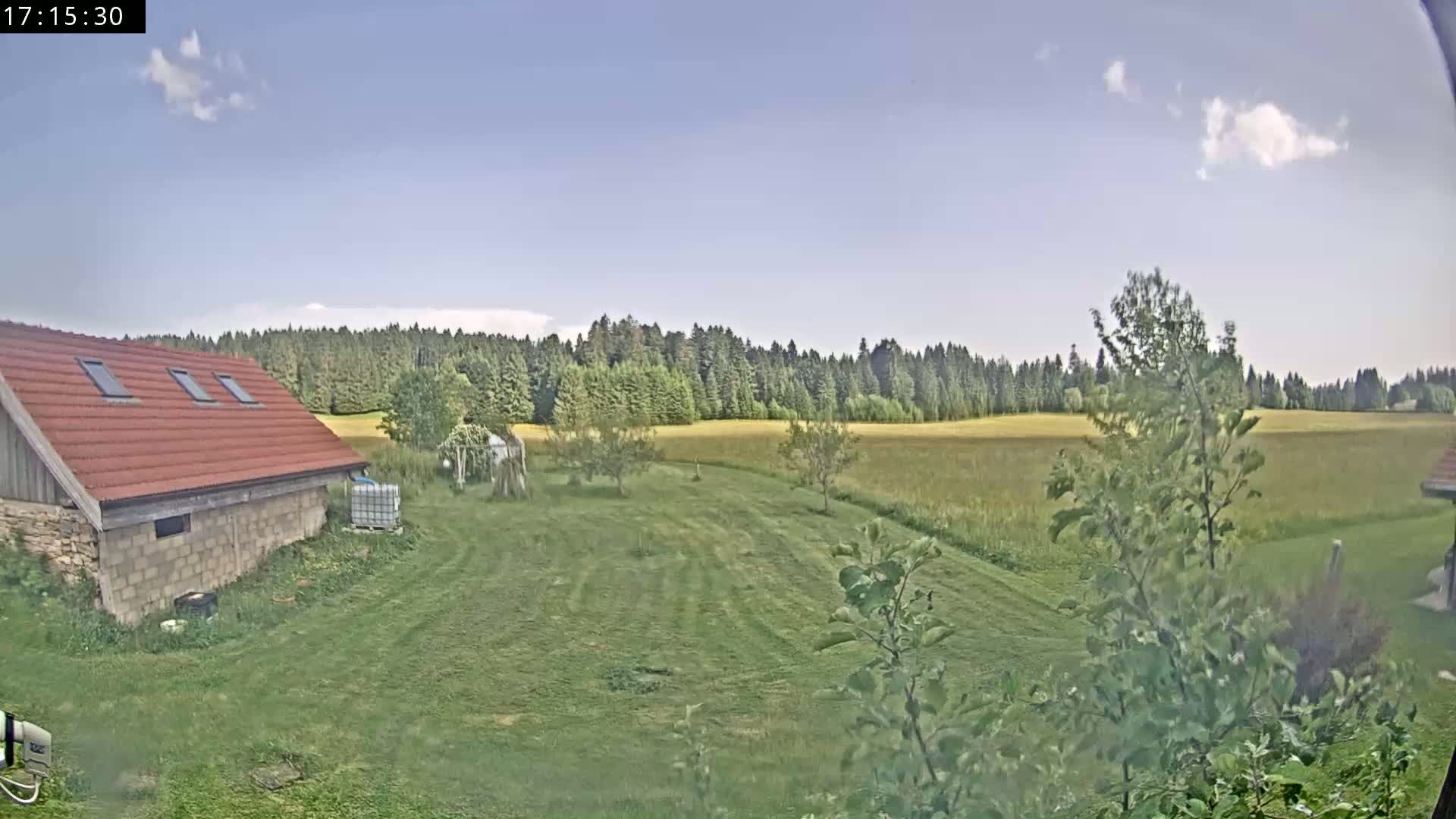 A red-roofed building sits on a grassy yard, overlooking a field and a line of trees under a partly cloudy sky.