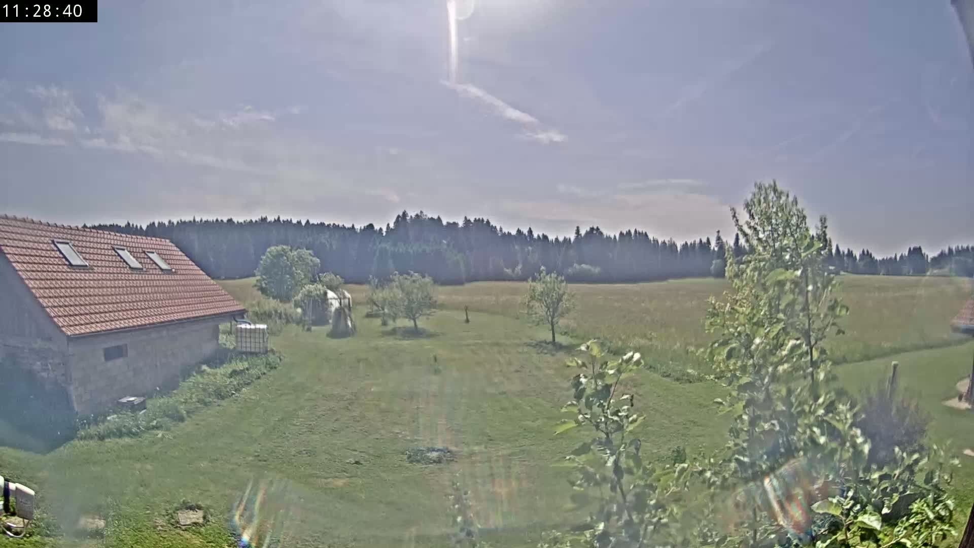 A stone building with a red tile roof sits on a grassy yard, overlooking a field and a distant forest under a partly sunny sky.