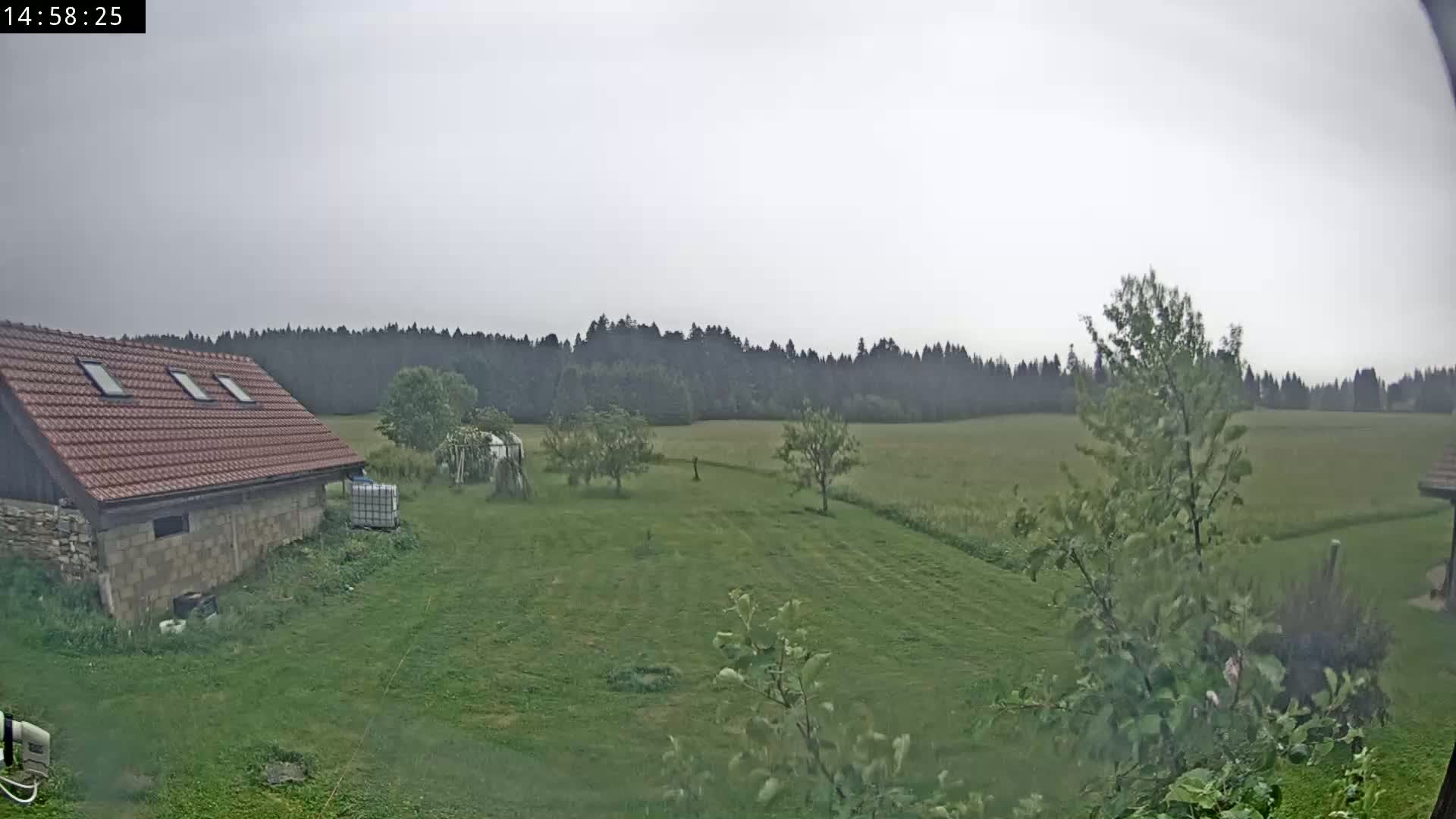 A stone and brick building with a red tile roof sits on a grassy yard overlooking a field and forest under an overcast sky.